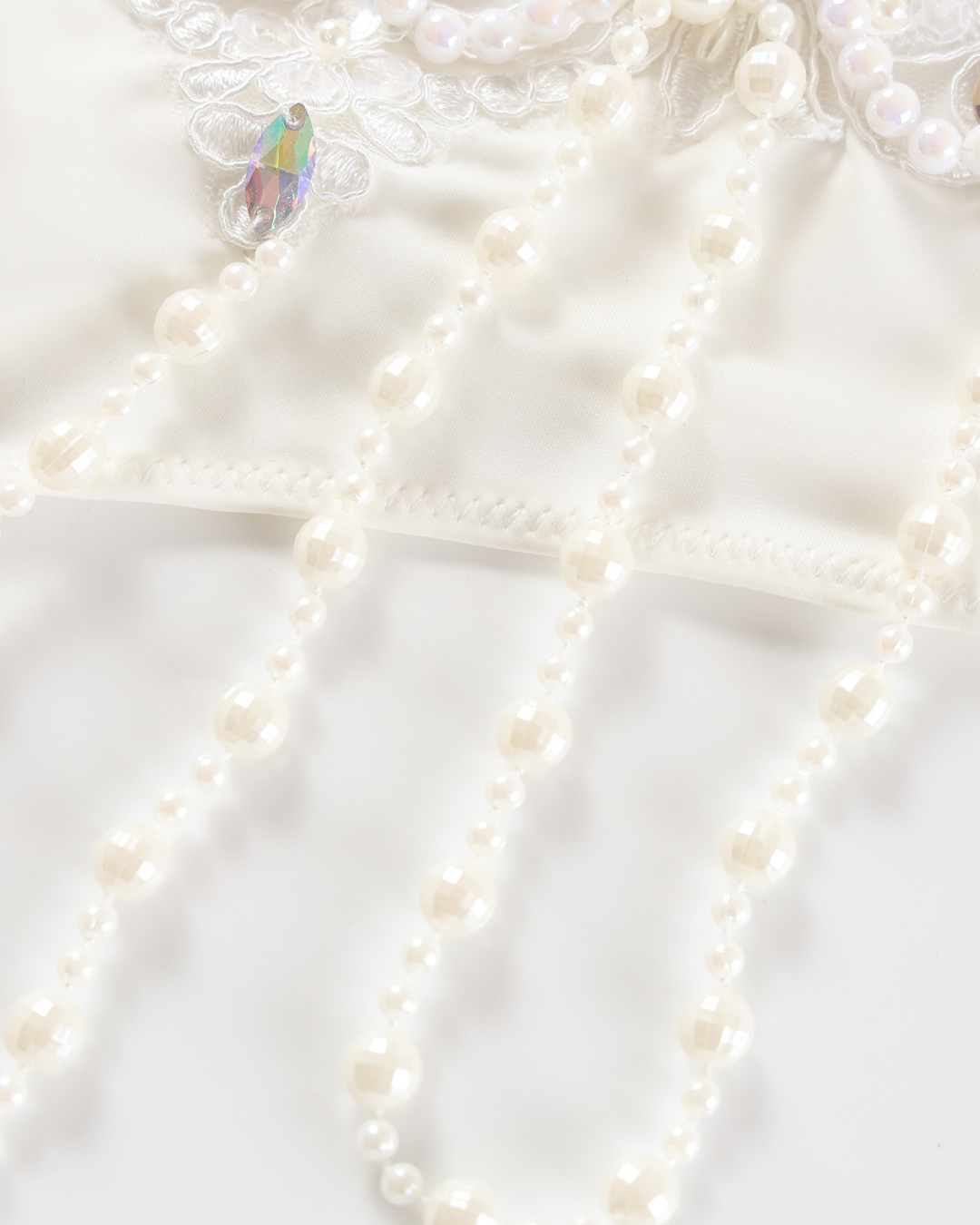 Close-up of Opheline - White - Bottom featuring imitation pearl beads on white fabric with lace embroidery and an iridescent teardrop-shaped gem.