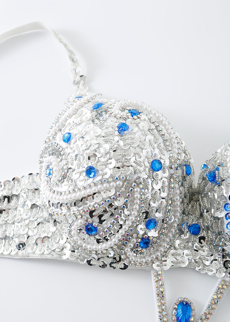 A close-up of the Imperya - Silver - Top, a bra embellished with silver sequins, rhinestones, and blue gems on a white surface—ideal for ChillFit Rave or as standout rave festival wear.