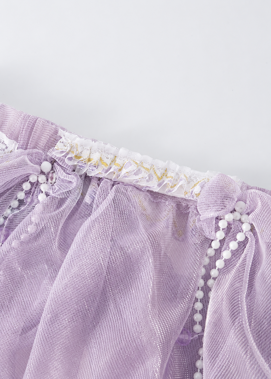 Close-up of the Gossamer-Purple-Skirt, made from lavender sheer fabric with white lace trim, gold accents, and decorative white bead garlands, displayed against a plain white background—an ideal option for festival or rave wear.