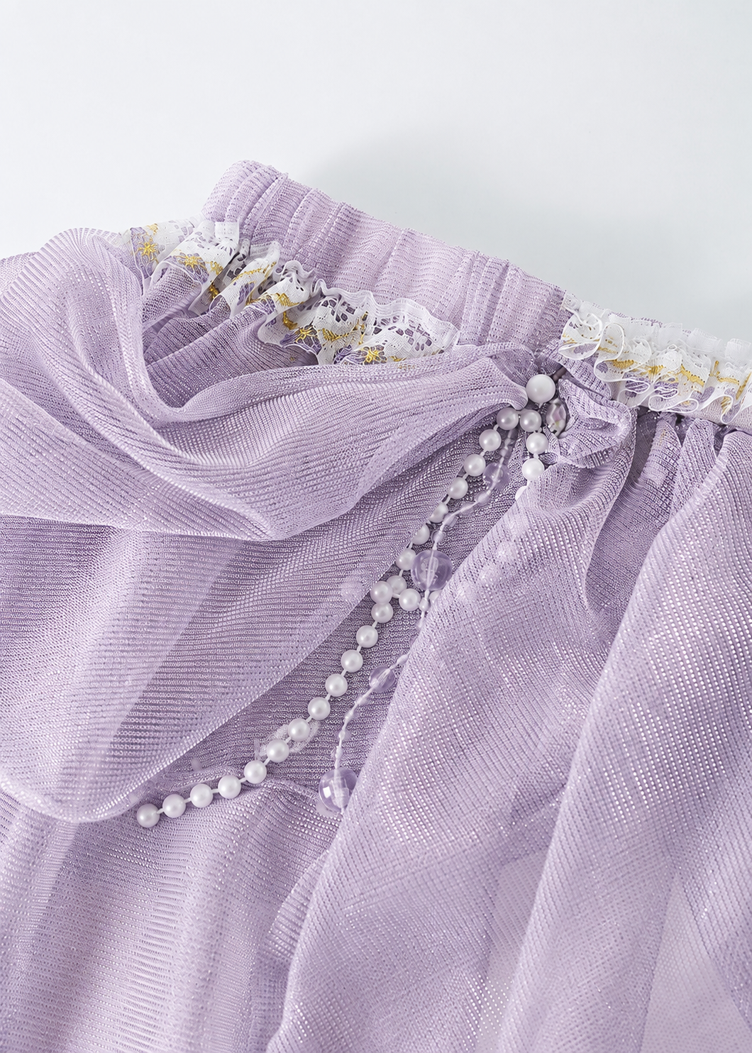 Close-up of the Gossamer-Purple-Skirt, ideal for festival or rave outfits, showcasing light purple sheer fabric accented with white lace trim, gold detailing, and a strand of white pearls draped elegantly across the gathered waistband.