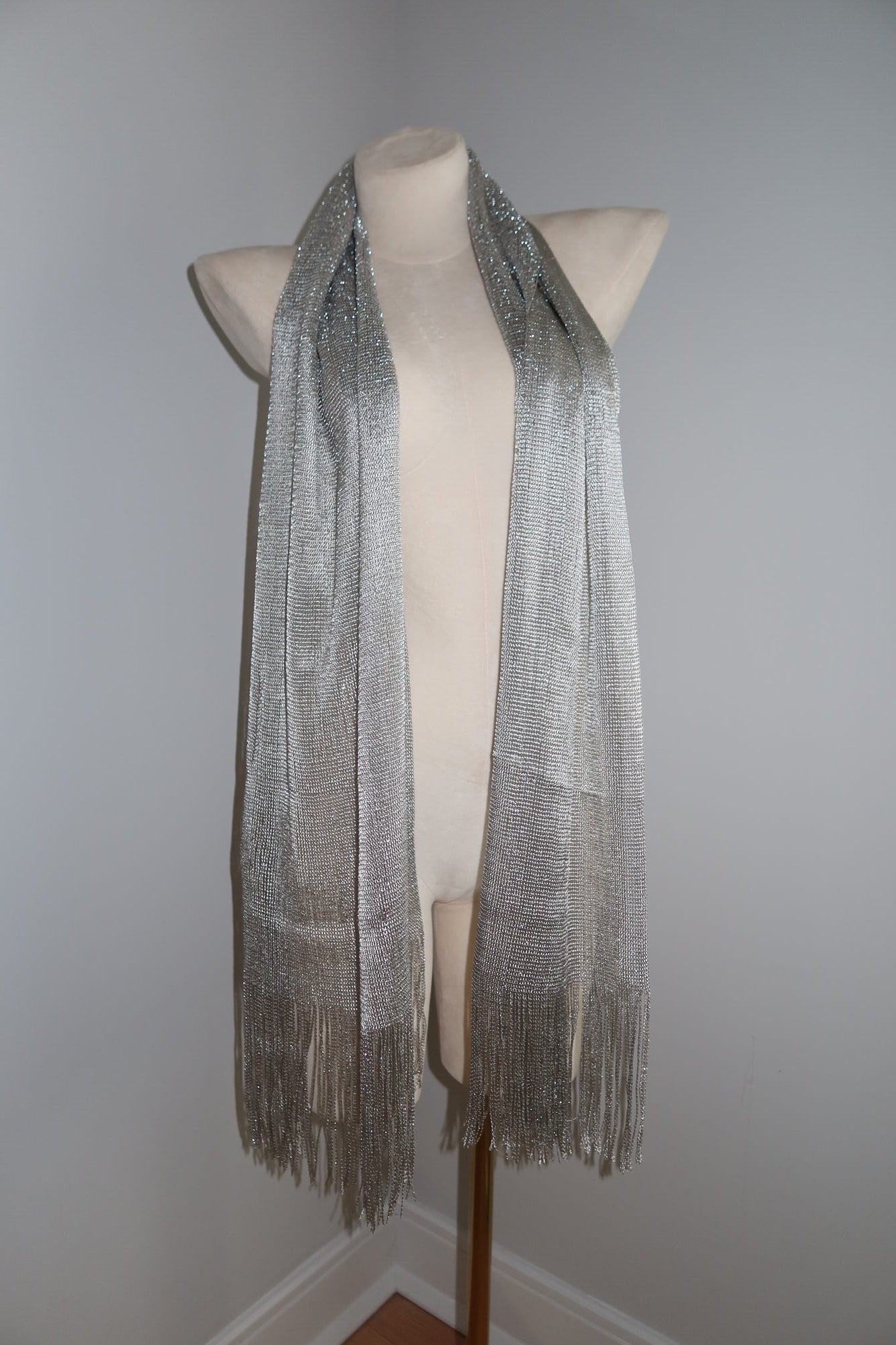 A mannequin showcases the Pashmina - Silver Sheer Fringe, a long, shimmery festival wrap with fringed ends, against a plain light wall—ideal for elevating your festival look.