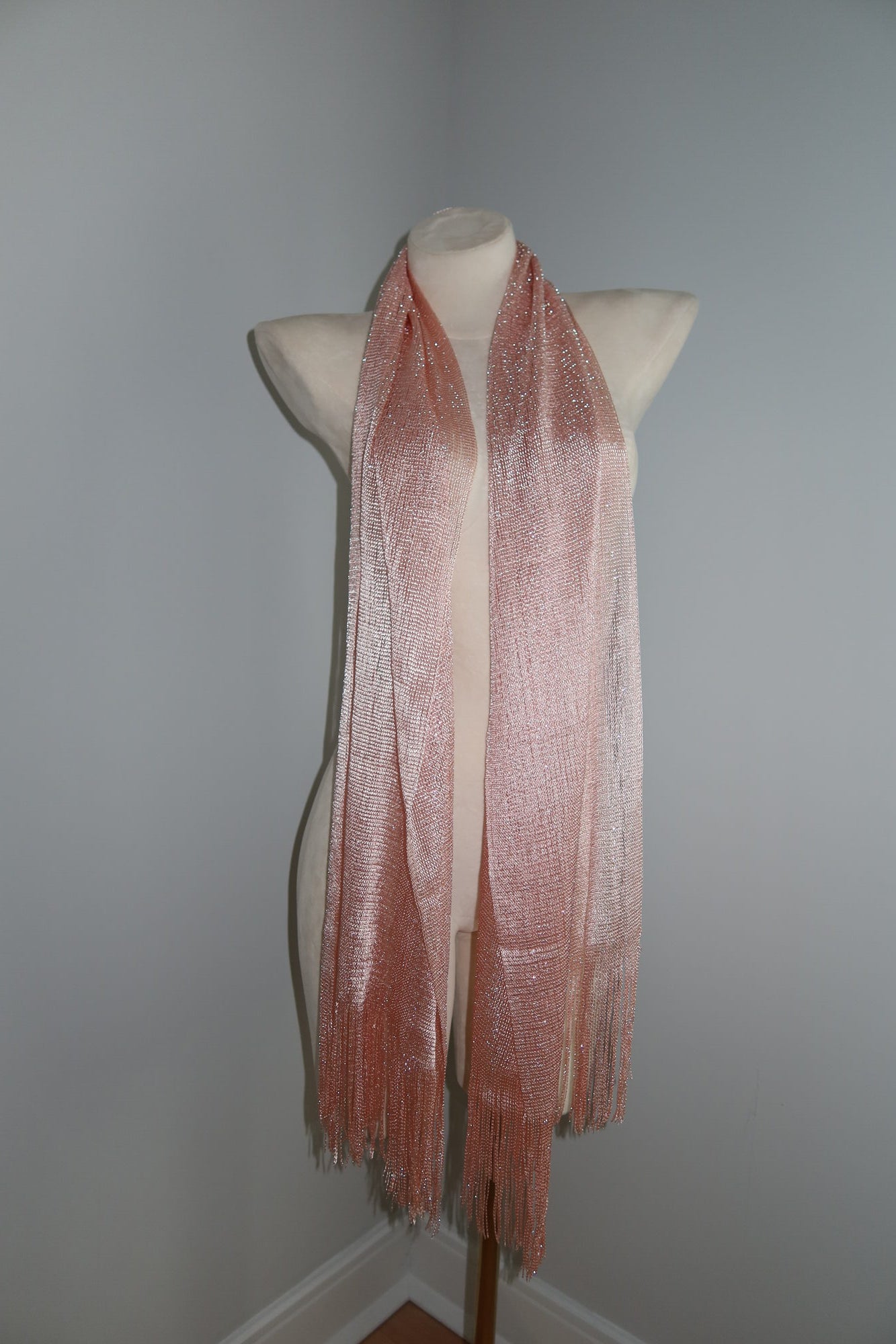 The Pashmina - Blush Sheer Fringe is a pale pink, shimmery scarf with fringed ends shown on a beige mannequin torso against a gray wall—ideal as a chic wrap for music festivals and festival fashion.
