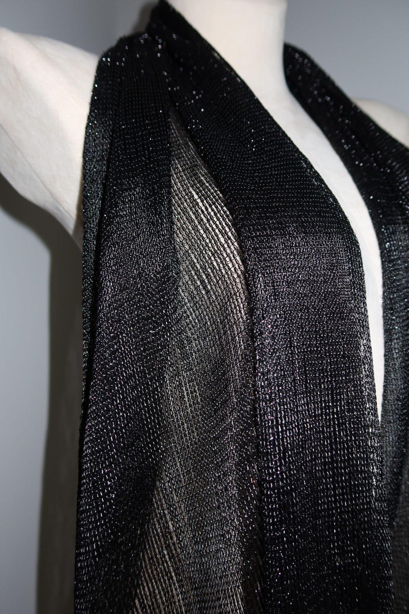 A close-up of a mannequin showcases the Pashmina - Black Sheer Fringe, its shiny, semi-sheer fabric draped stylishly as a festival wrap, highlighting the texture and shimmer against a plain background.