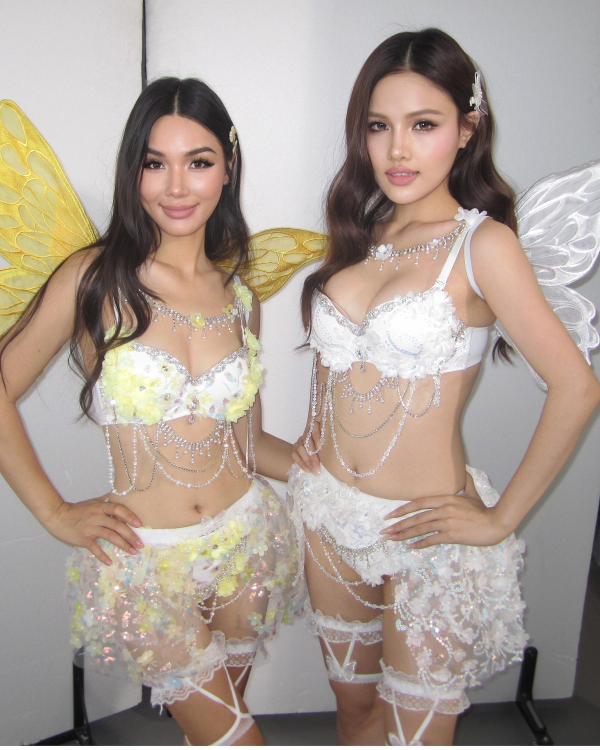 Two women pose together in dazzling festival outfits, each wearing ornate white costumes with sheer skirts and intricate beaded details. One features yellow wings, the other white. Both showcase the stunning Lavendelle - White - Top with delicate floral sequin appliques and detailed rave bra designs, standing against a plain background.