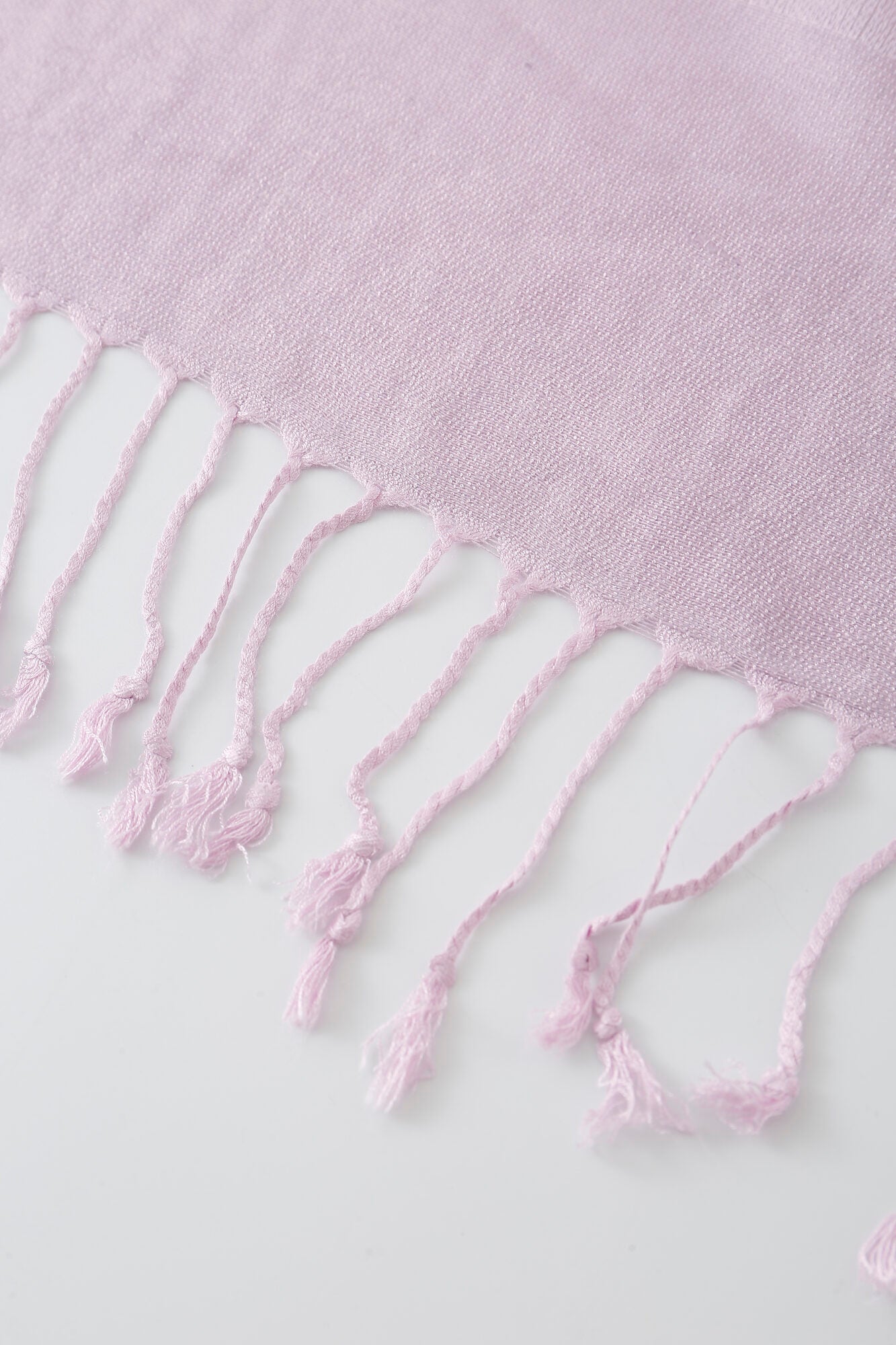 A close-up of the Pashmina - Lavender Solid Fringe, featuring light lavender woven fabric with fringed tassels along the edge, laid flat on a white surface.