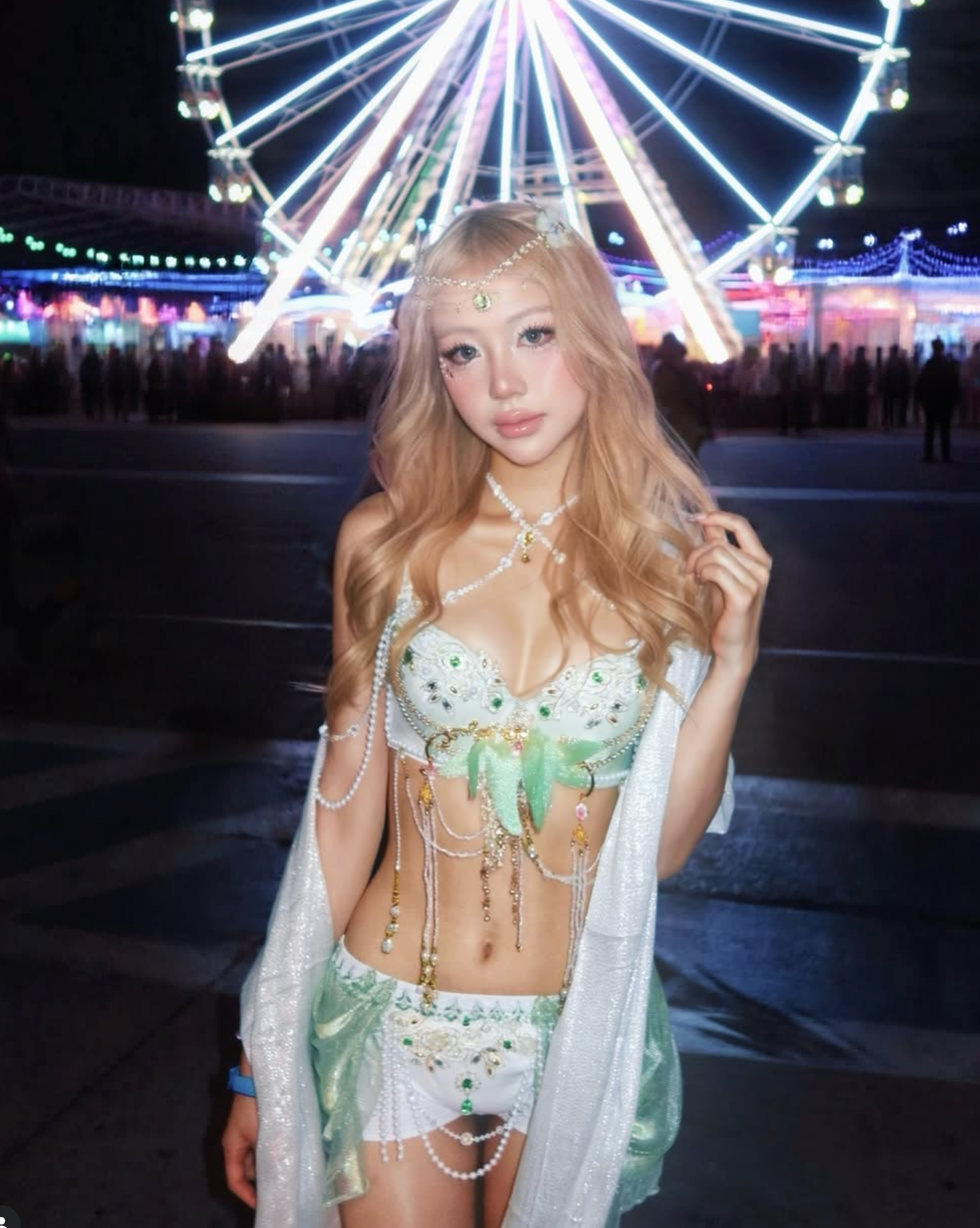 A woman in festival fashion poses in a beaded outfit and the Silver Sheer Fringe Pashmina, framed by a brightly lit Ferris wheel at night.
