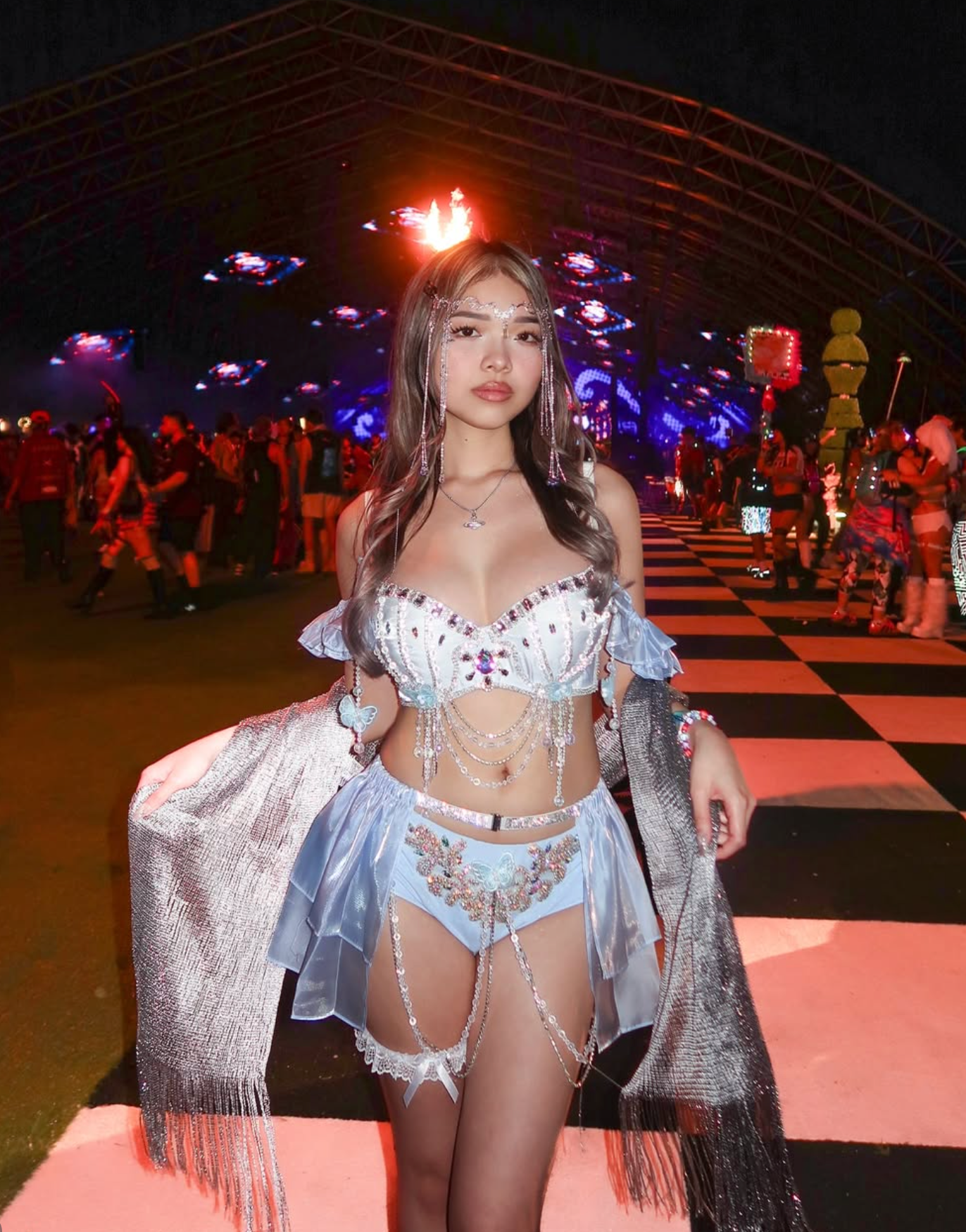 A woman wearing the Pearl Wing - Blue - Bottom poses on a black-and-white checkered walkway at a nighttime outdoor event, surrounded by colorful lights and people.