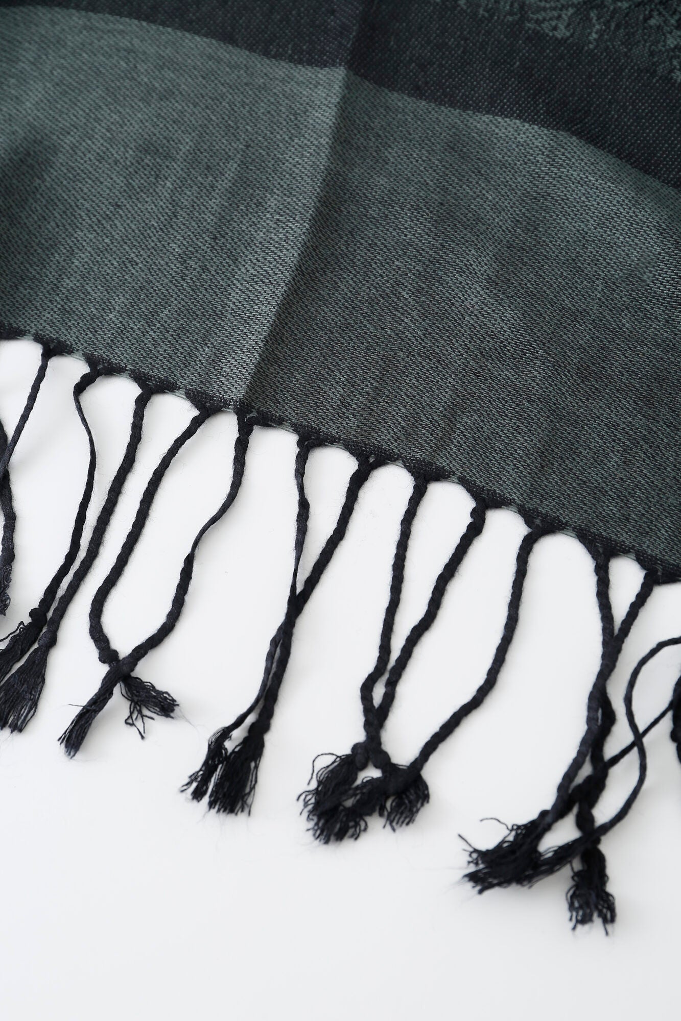 Close-up of the edge of the Pashmina - Slate Paisley Fringe scarf in dark green and black, featuring several twisted tassels hanging on a white surface.