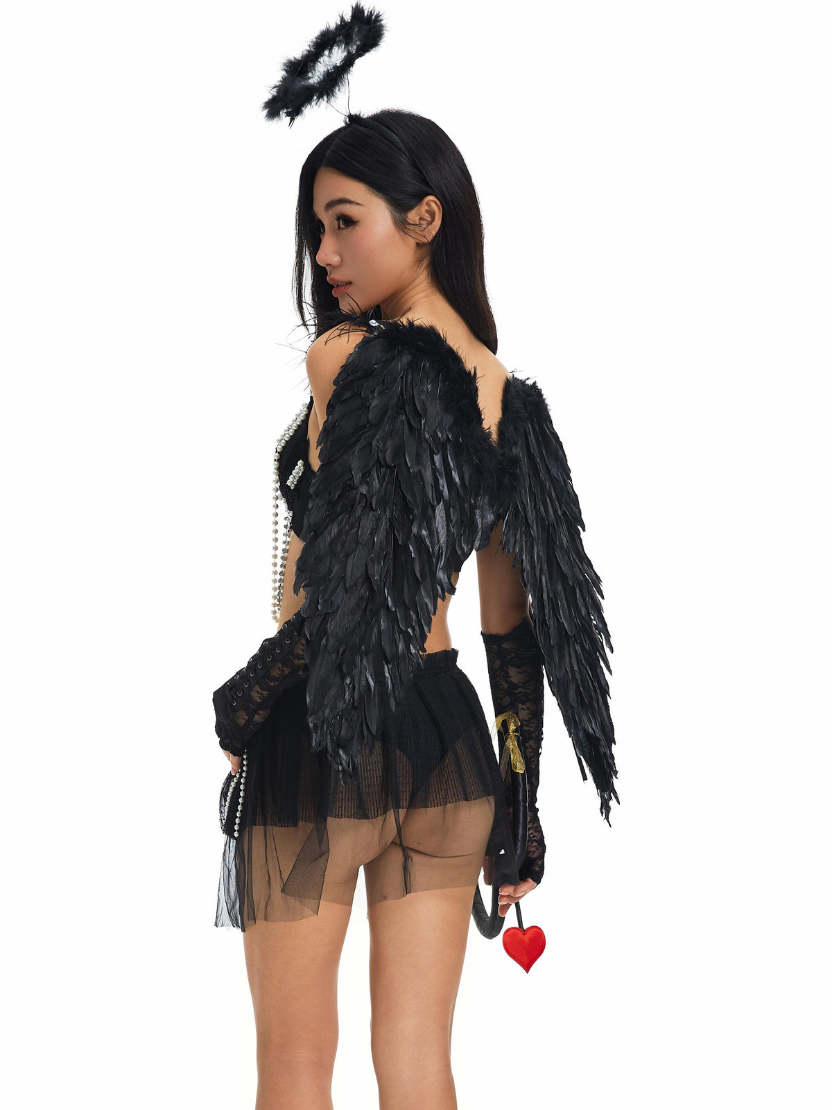 A woman wearing a black angel costume featuring Devil Wings- Black, a dramatic halo headband, lace gloves, a sheer skirt, and holding a red heart-shaped Halloween accessory stands against a plain white background.