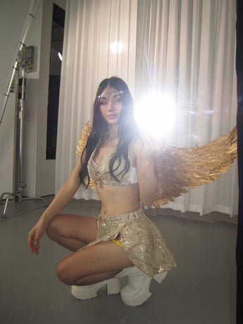 Dressed in The Divine- Selene Gold costume with shimmering gold angel wings, the person squats indoors to take a mirror selfie, their face hidden by the camera flash.