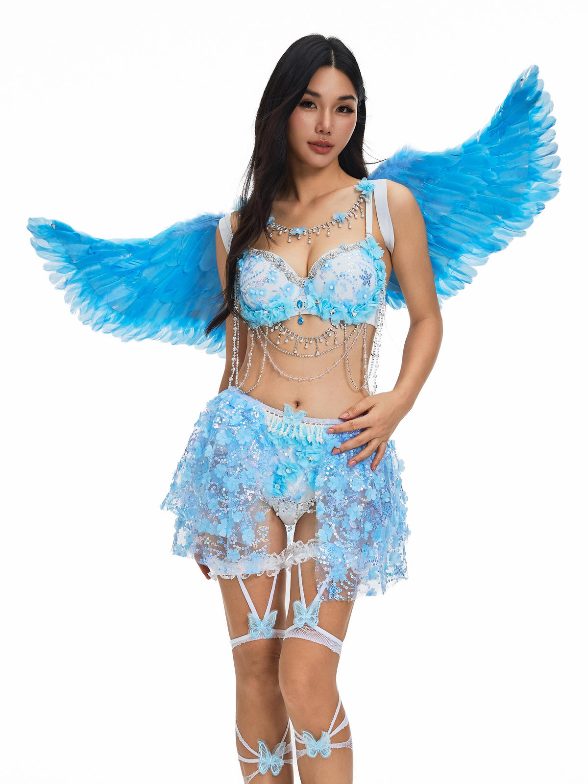 A woman wearing The Fae- Lavendelle Blue—a light blue, jeweled costume featuring blue angel wings, blue butterfly thigh-high stockings, and a lace skirt—poses against a white background.