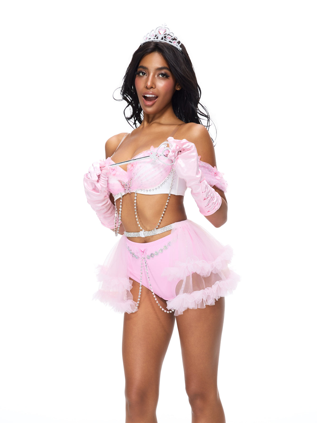 A woman is wearing the Princess 2 Pieces Set- Pink, featuring a tiara, wand, gloves, pearl necklace, and ruffled skirt, as she poses against a plain white background.