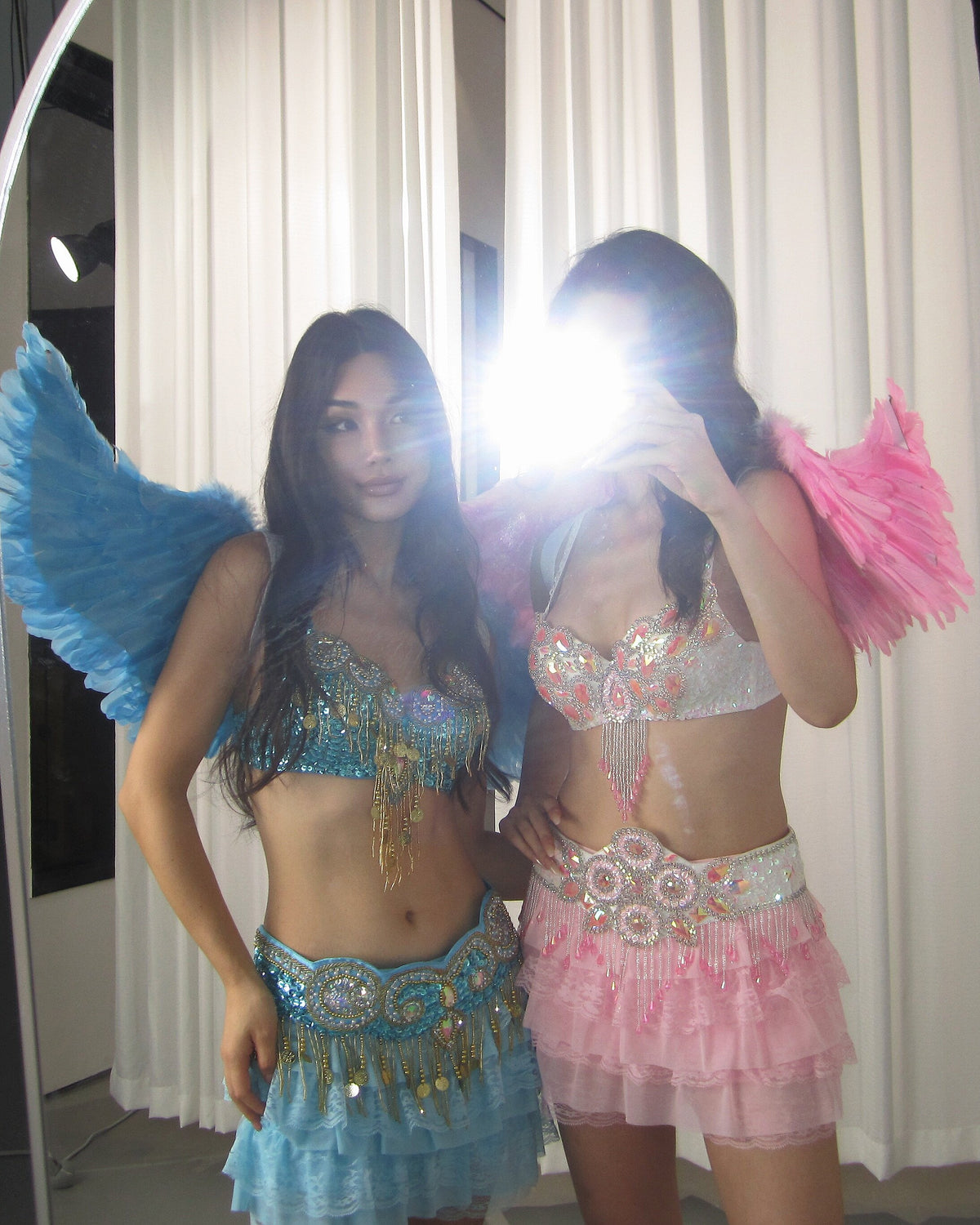 Two women pose for a mirror selfie in The Runway Angel- Notics Aqua costumes, their shimmering angel wings on display—one face partially obscured by the camera flash.