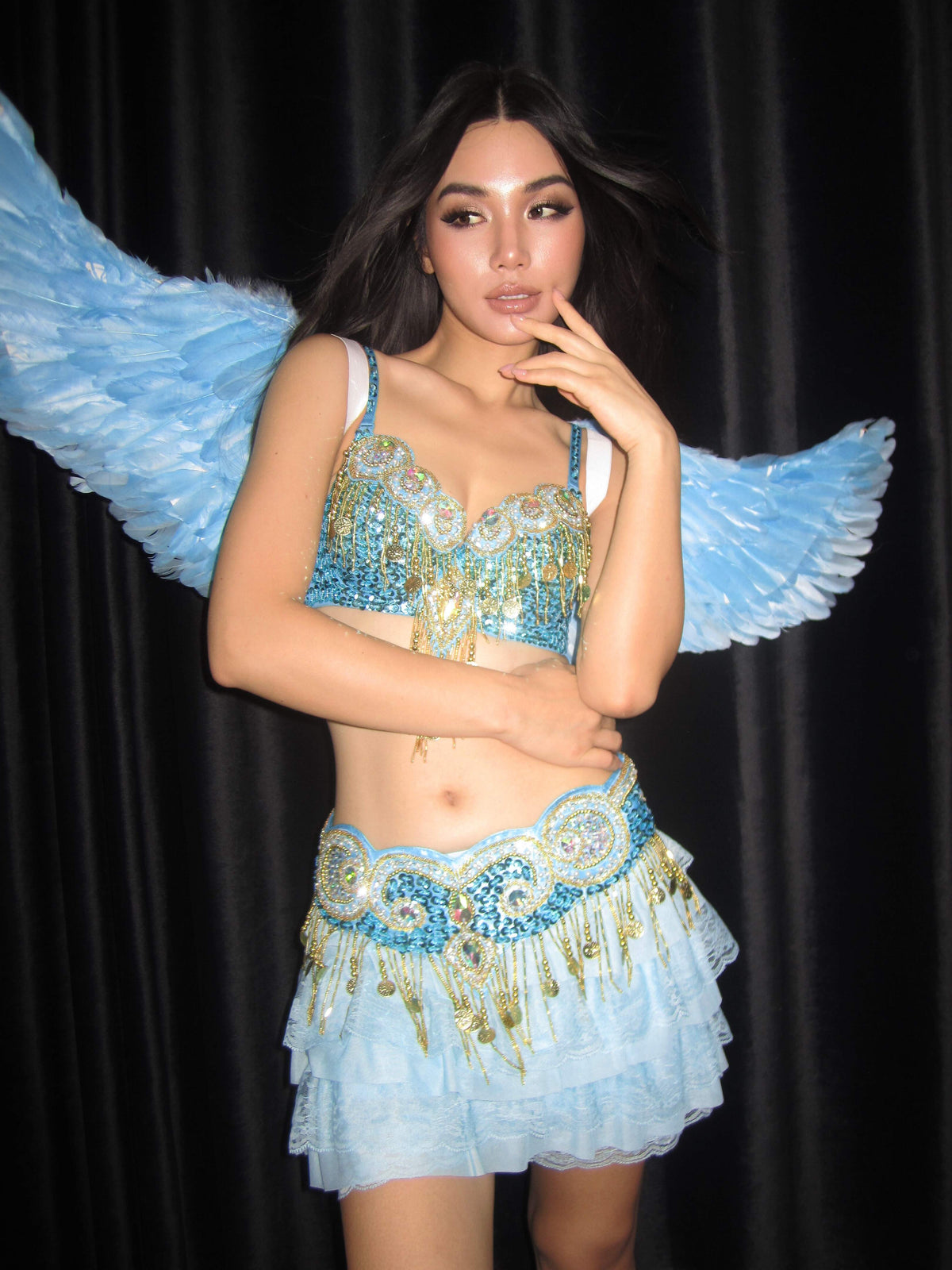 A person wearing a sparkling blue and gold Halloween costume with a layered skirt and matching oversized Angel Wings- Blue poses in front of a black curtain.