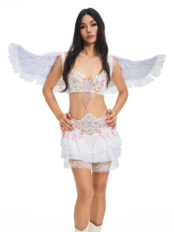 A woman stands against a plain white background, wearing “The Runway Angel- Vixen White” ensemble, featuring striking angel wings, a beaded bra, a layered skirt, and white boots.