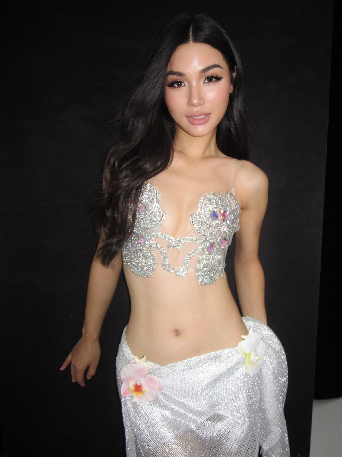 A woman wearing The Siren- Platinum Tassel Aurora, a silver jeweled bra top and matching skirt adorned with flowers, elegantly drapes a White Sheer Fringe Pashmina over her shoulders as she poses in front of a black background.
