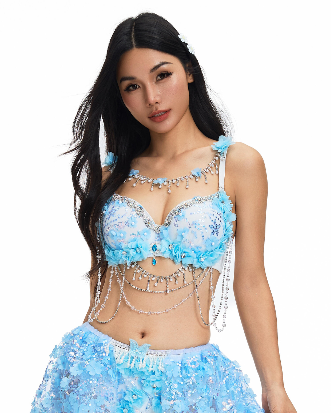 A woman poses against a white background wearing the Lavendelle-Blue-Top, an eye-catching festival outfit featuring a light blue embellished bra with floral and crystal details, silver chains, and a matching skirt.