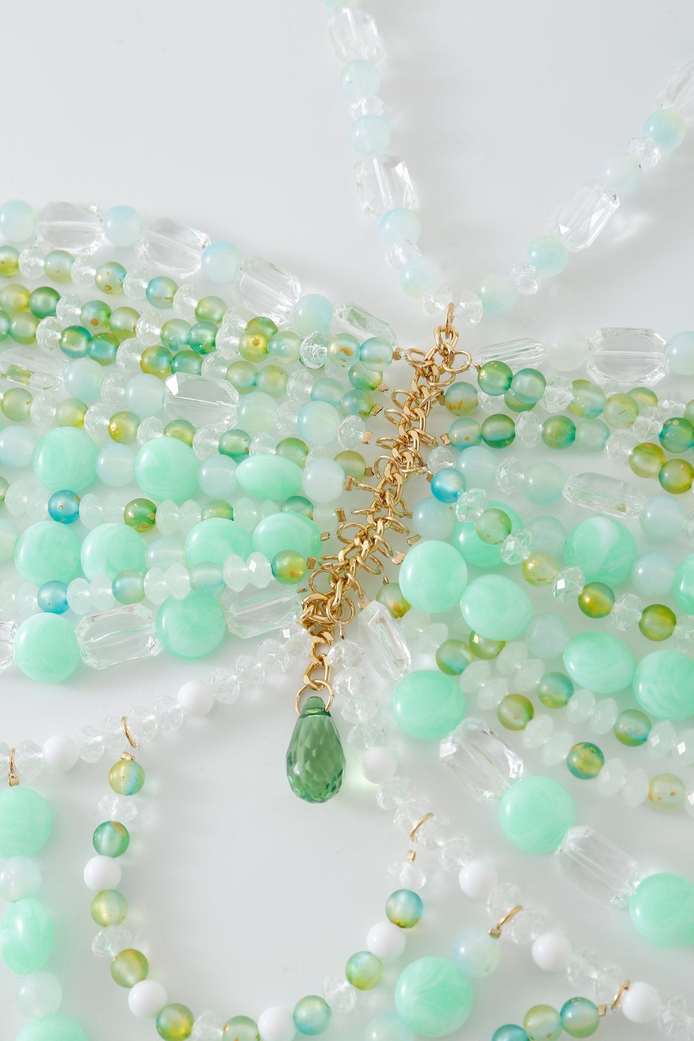 Several strands of green, white, and clear beaded necklaces are arranged on a white surface, reminiscent of accessories that would perfectly complement the Anemone w Mini Skirt- Full Set. A gold chain with a striking green teardrop pendant sits elegantly in the center.