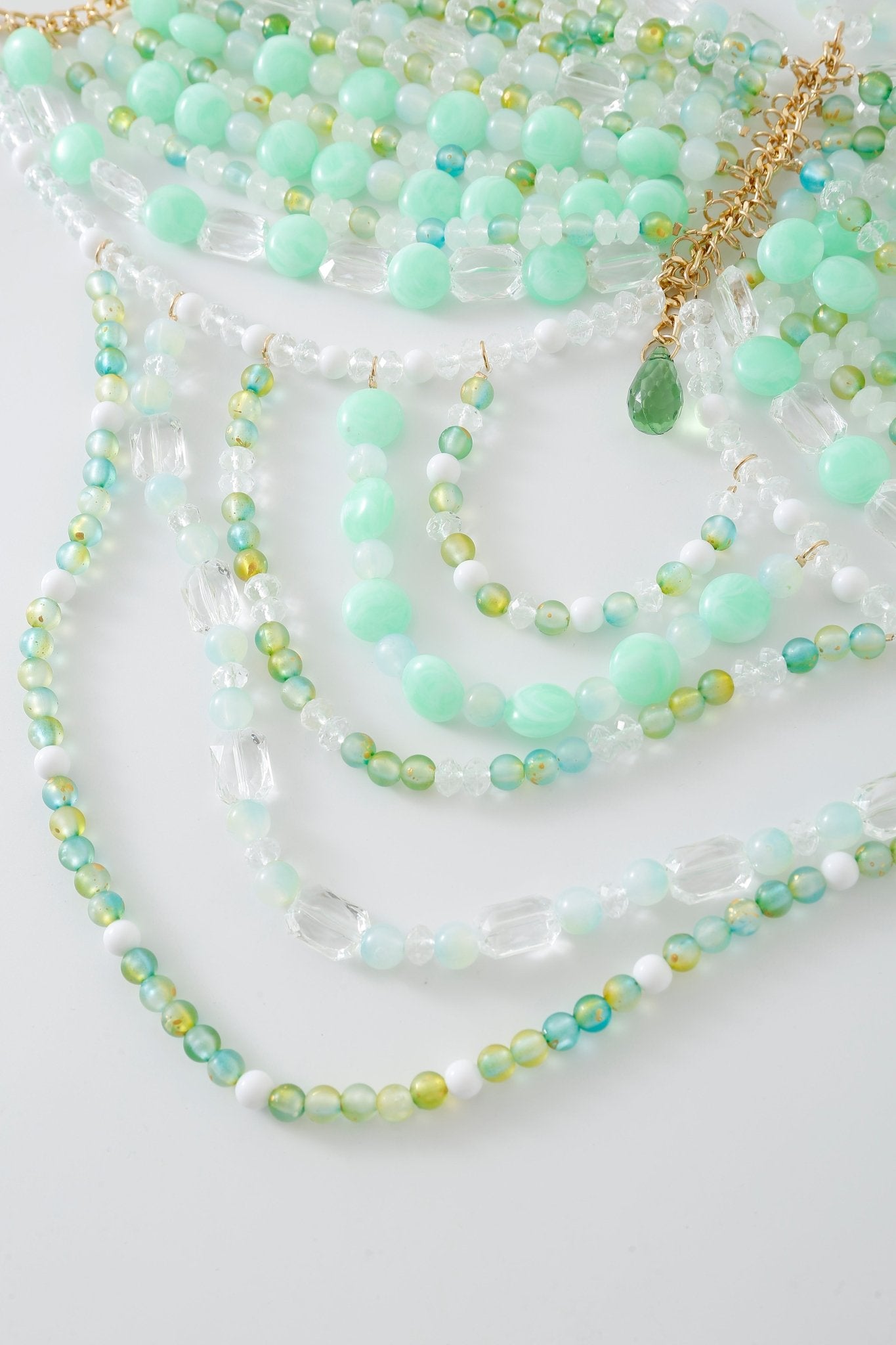 The Anemone w Pashmina- Full Set features several beaded necklaces in shades of green, blue, and white with gold accents, arranged on a white surface and evoking the intricate detailing of a beaded top.