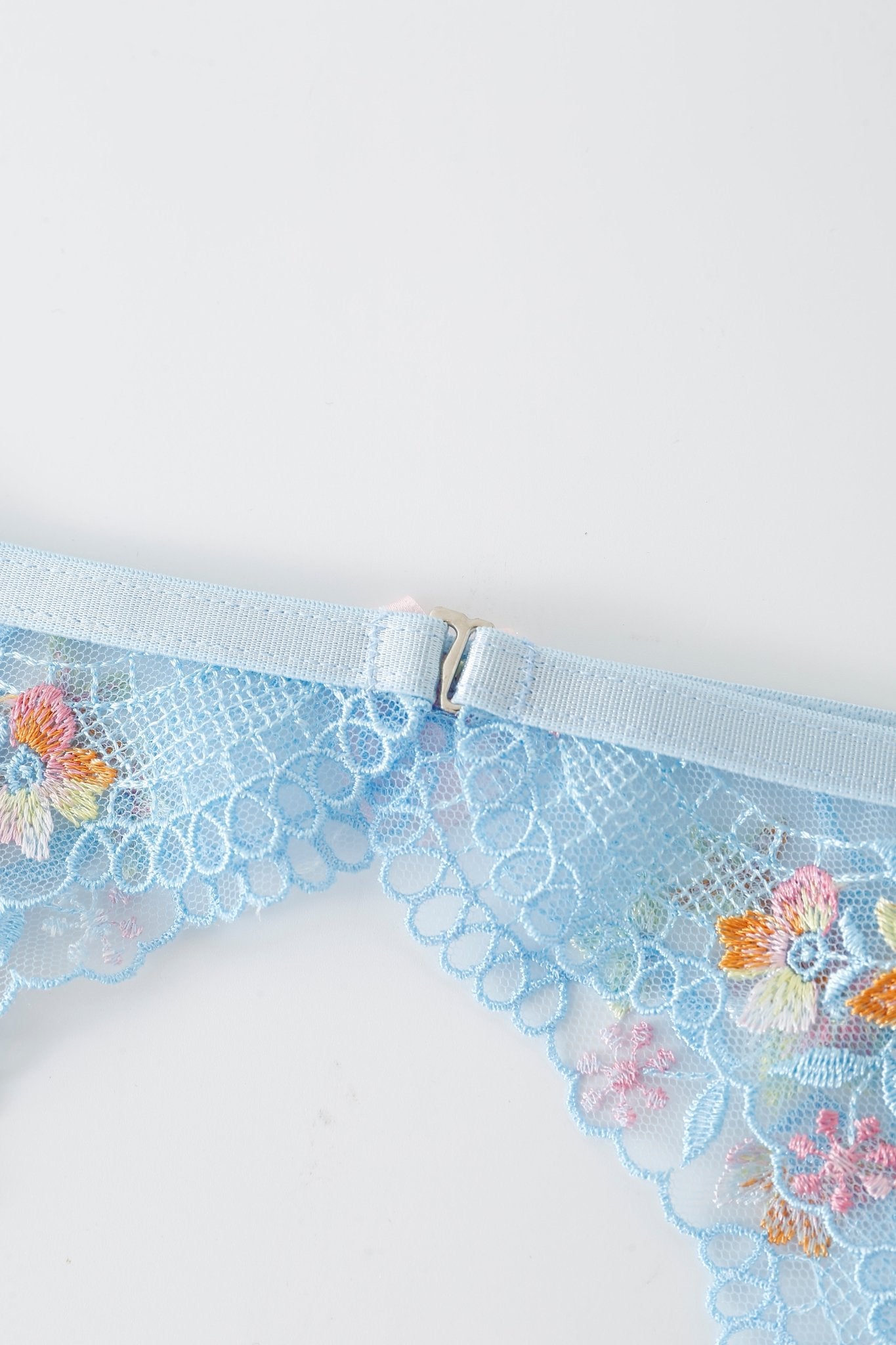 Close-up of the Azura w Mini Skirt - Full Set featuring light blue lace fabric with floral embroidery, a small metal hook clasp, and delicate mesh texture reminiscent of fine lingerie. Subtle rhinestone detailing adds an extra touch of elegance against the white background.
