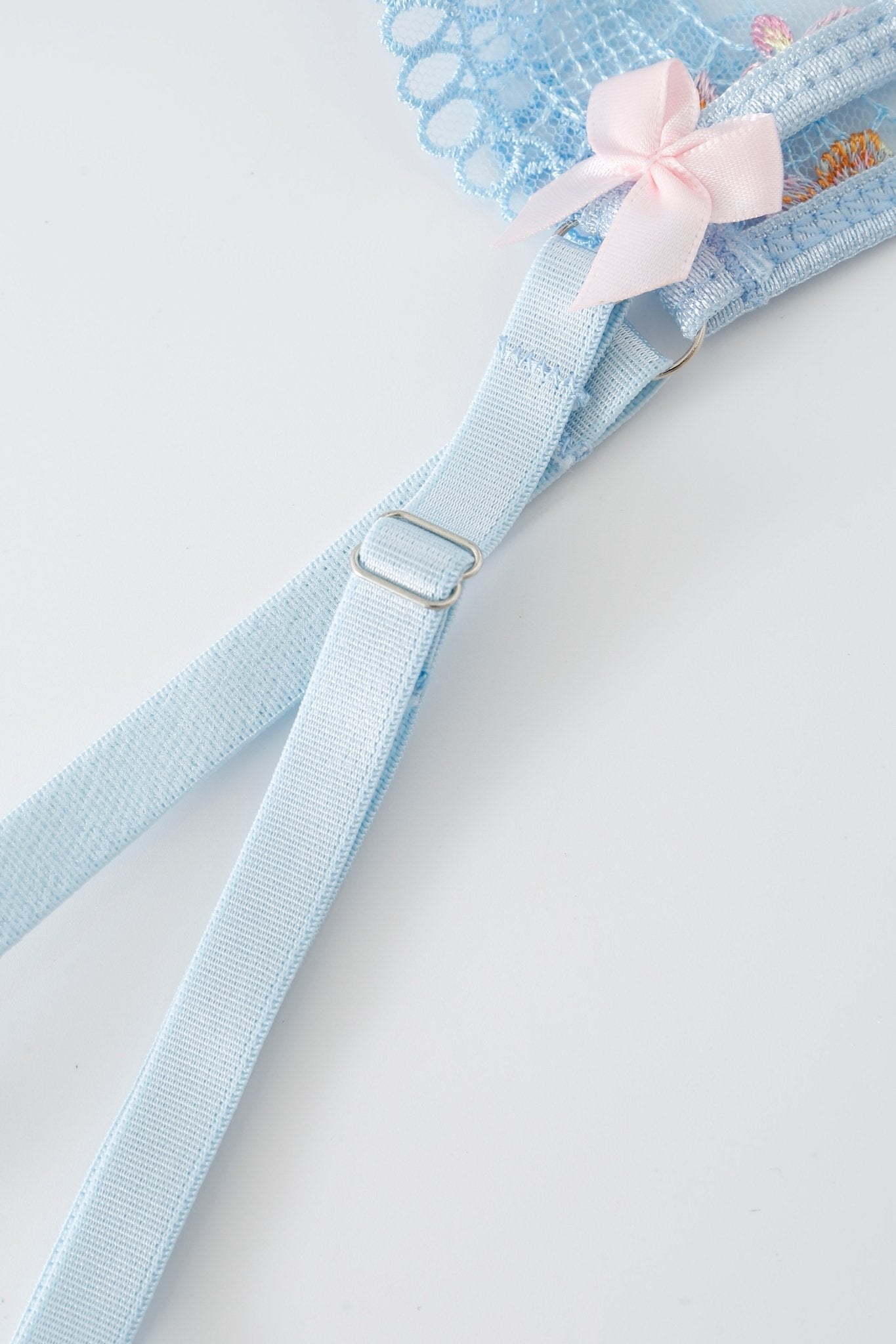 Close-up of a light blue strap from the Azura w Mini Skirt- Full Set, featuring an adjustable metal slider and a small pink bow on lace fabric, set against a plain white background.