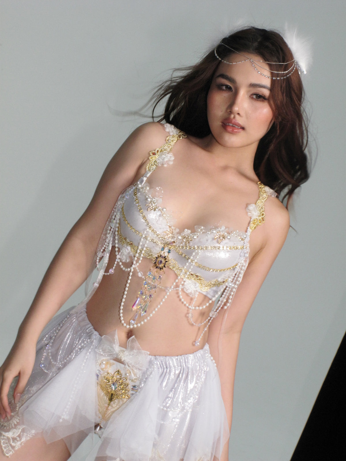 A woman stands against a plain background, showcasing the CROWN - Top: an elegant, handmade festival bra featuring a white, pearl-adorned costume with gold accents, a sheer skirt, and a coordinating headpiece.