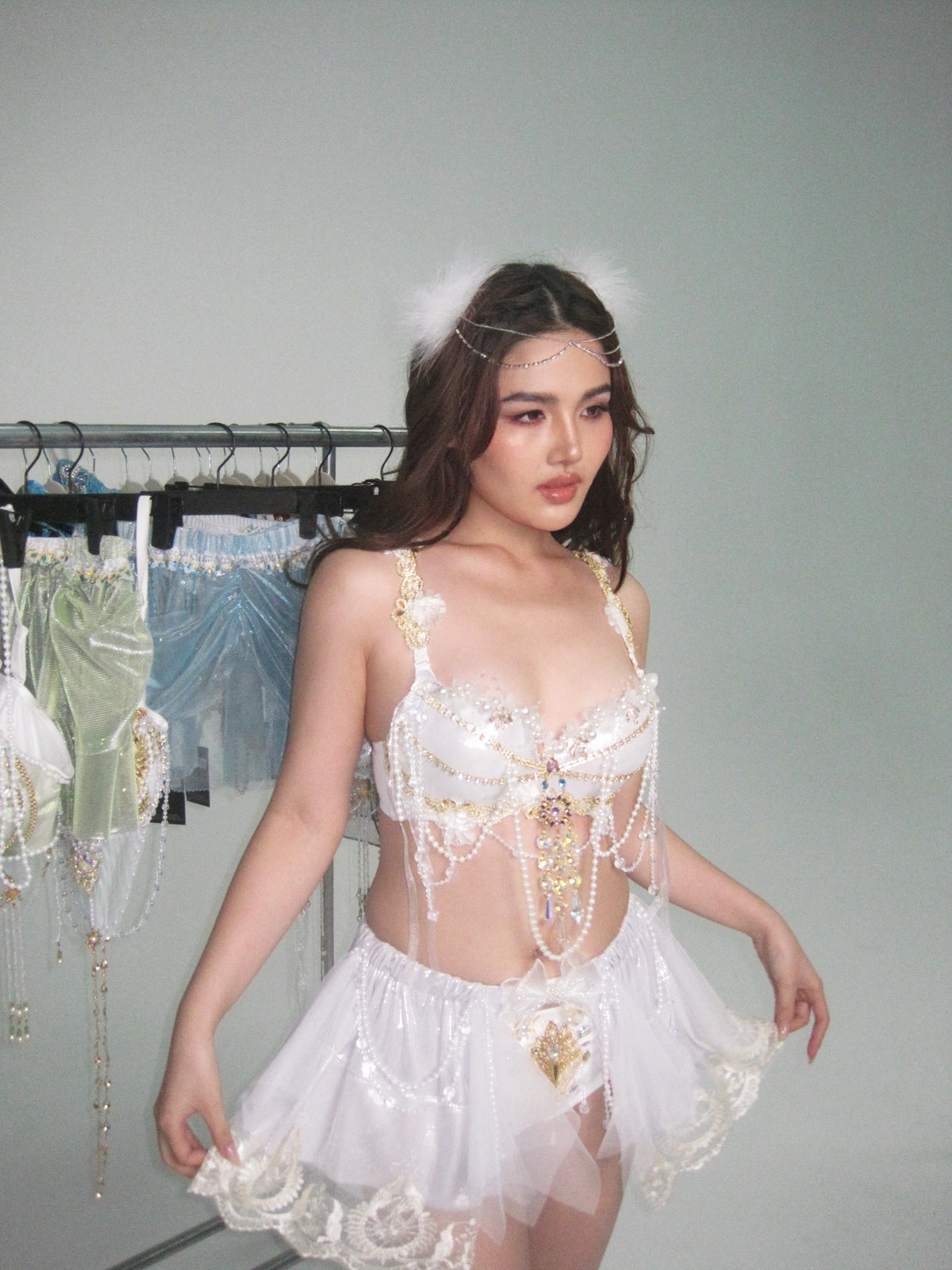 A person wearing the CROWN - Skirt, featuring delicate beading, lace details, and a flowing tulle design, stands in a studio; festival-ready clothing items hang on a rack in the background.