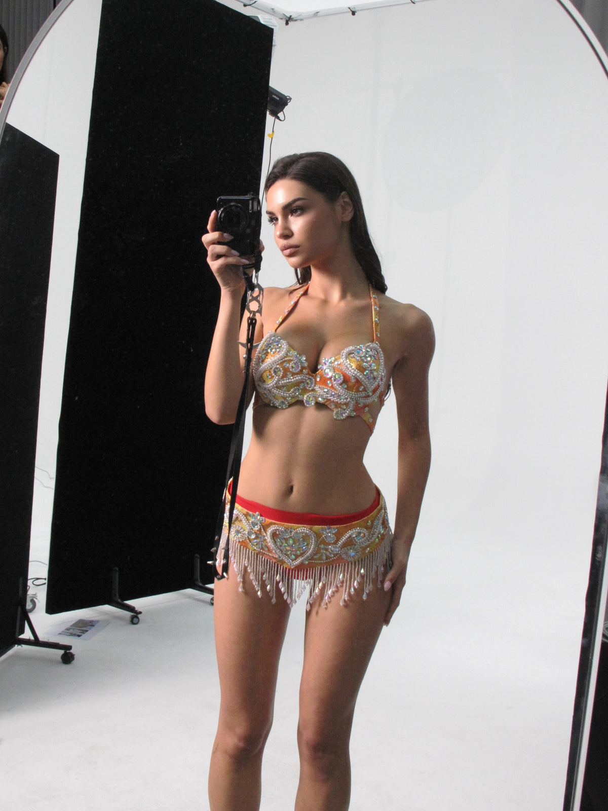 Wearing the Domina - Coral - Top, a woman takes a mirror selfie in a photography studio with lighting equipment visible, showcasing this paisley-patterned, handcrafted bikini top with fringe—perfect for festival wear.