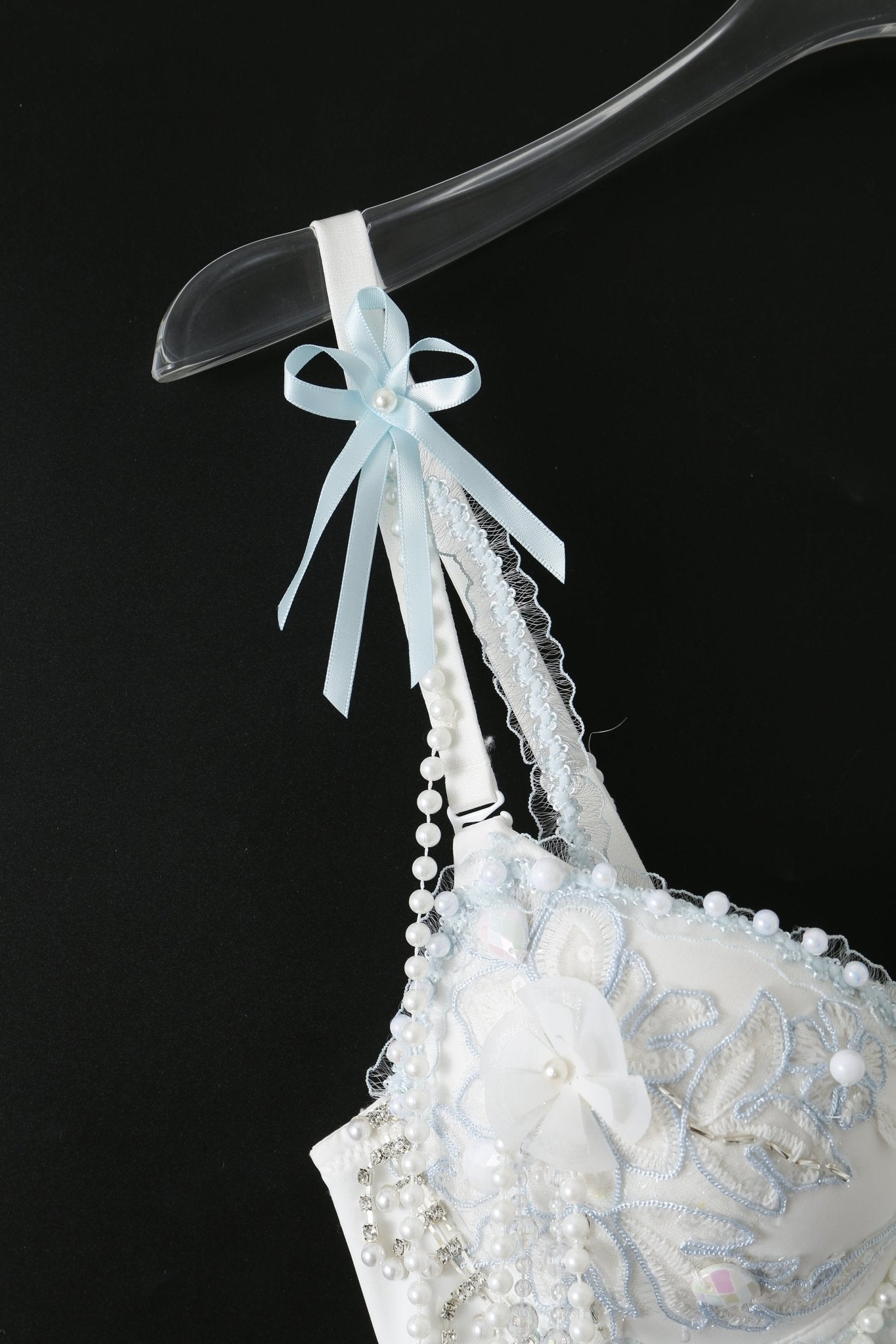 Eira - Full Set - Blue: White decorative bra featuring embroidered florals, pearl accents, and a blue ribbon bow—perfect for raves—shown on a clear hanger against a black background.
