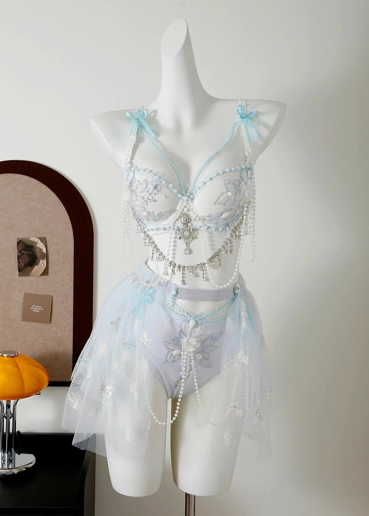 A white mannequin showcases the Eira - Full Set - Blue, a delicate light blue lingerie set with lace, sheer tulle, floral embroidery, and pearl accents—ideal handcrafted rave outfit inspiration—in a minimalist room with a lamp and corkboard.