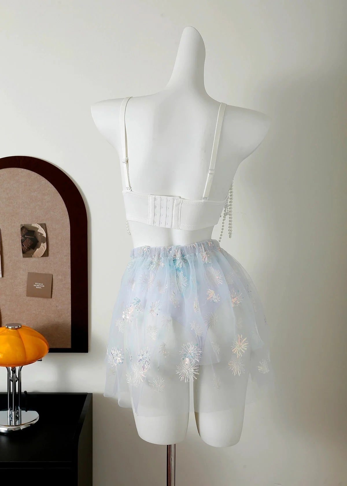 A white mannequin displays the "Eira - Full Set - Blue," a handcrafted rave outfit, in a minimal room featuring a small table lamp and wall art.