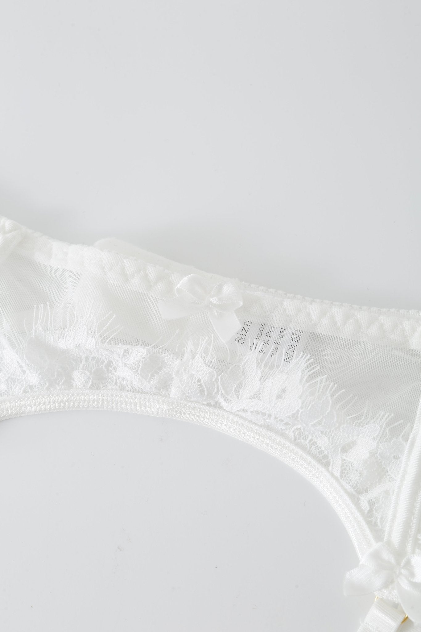 White lace underwear from the Eve w Mini Skirt - Full Set features a sheer mesh panel and small decorative bows, capturing the delicate charm of Eve lingerie as it rests on a plain white surface.