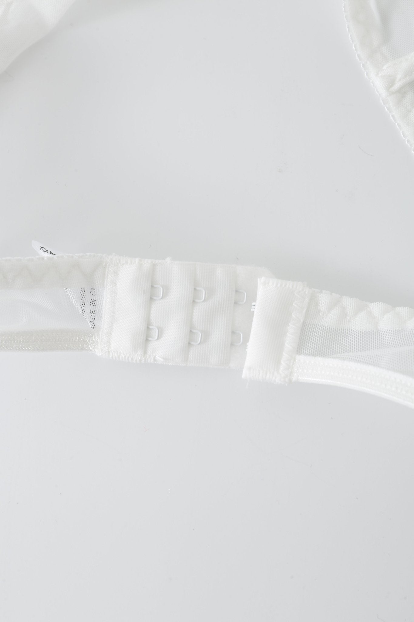 Close-up of a white bra strap from the “Eve w Pashmina- Full Set,” featuring a three-row hook-and-eye closure, lying on a white surface.