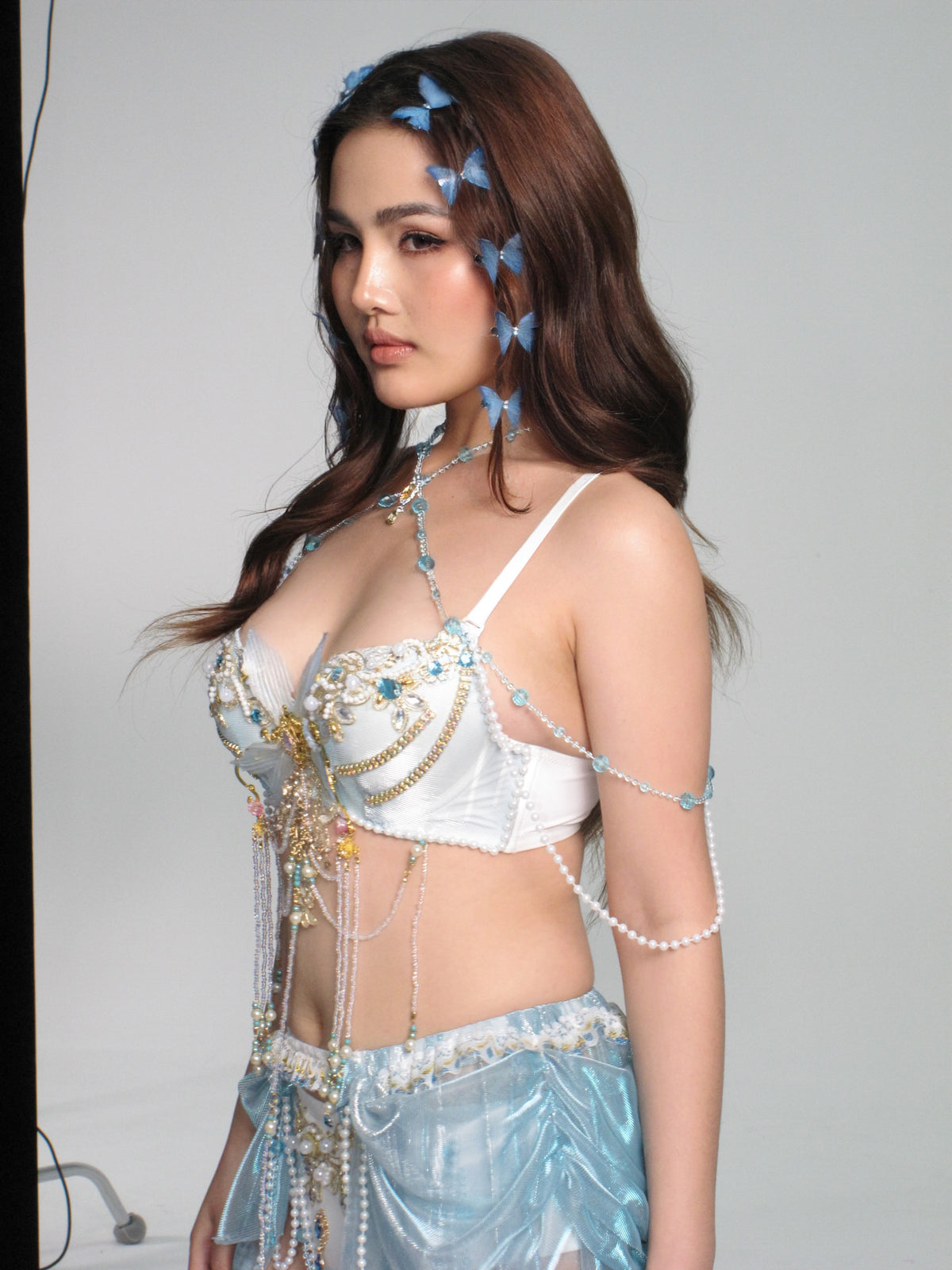 Wearing the GOSSAMER - Top - Blue, a woman with long brown hair styles her festival-ready look with beaded and pearl accents and blue butterfly accessories, standing against a plain background for an enchanting effect.