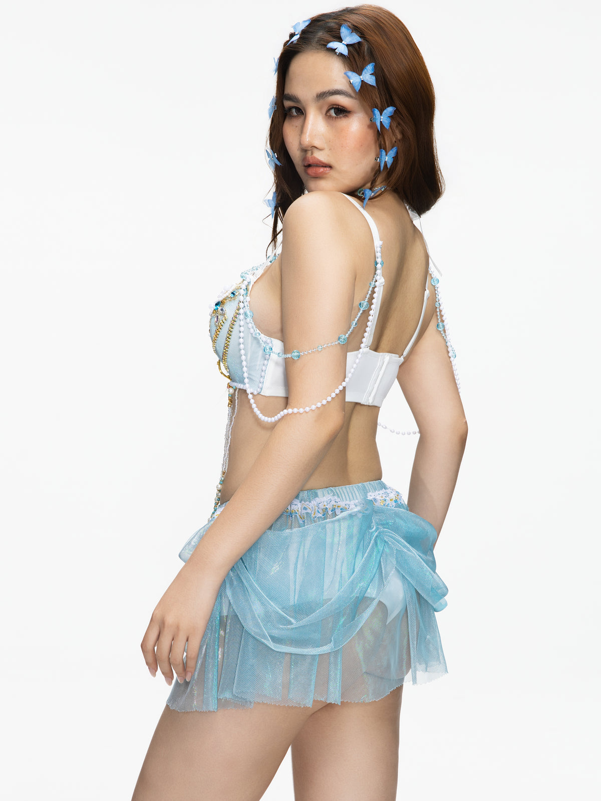 Wearing the GOSSAMER Top in Blue, a woman stands against a white background with butterfly hair accessories, looking over her shoulder.
