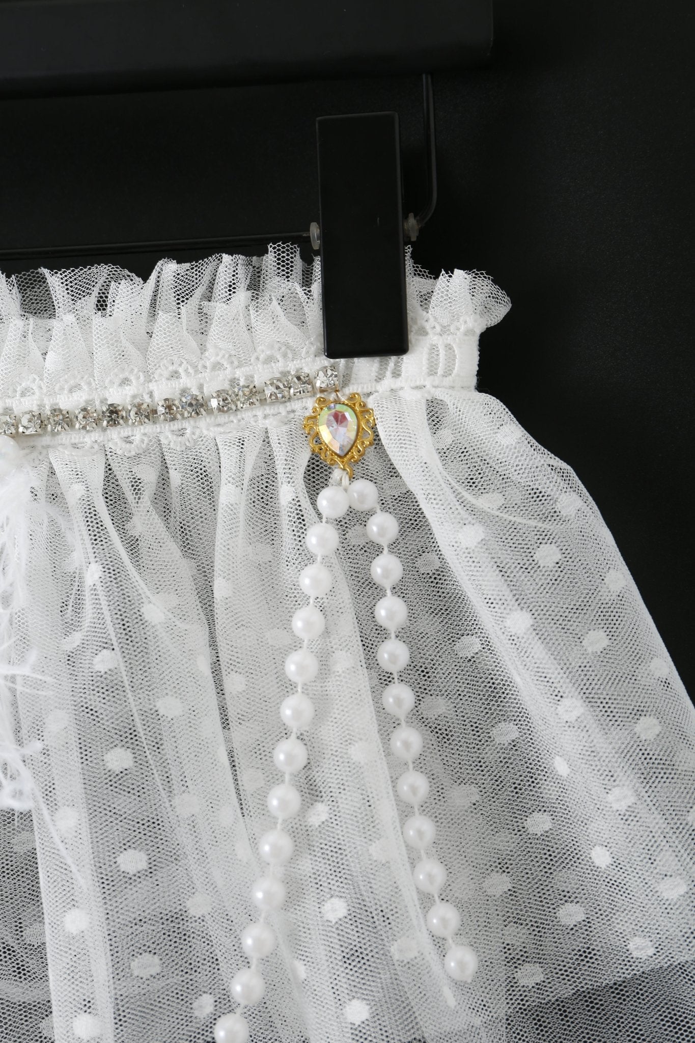 Close-up of the Haló - White - Skirt: white polka dot tulle with a pearl strand, rhinestone trim, and gold heart-shaped detail on a black hanger—stylish accents for your festival look or lace bodysuit.