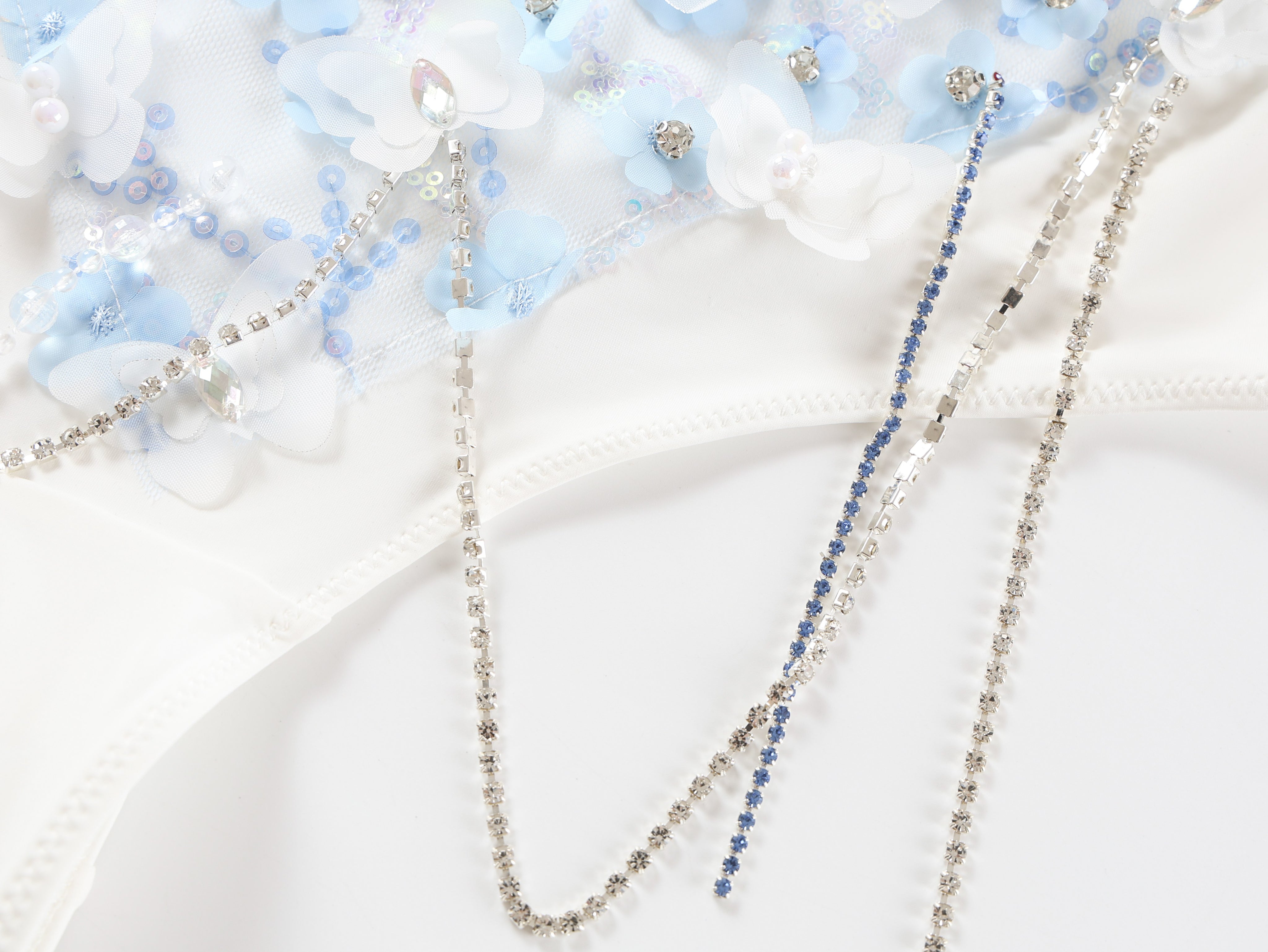 The Icemuse - Blue - Bottom features white fabric with light blue and white floral appliqué, bead accents, sequin floral hem, and three jeweled silver and blue chain strands—ideal for standout handmade rave outfits.