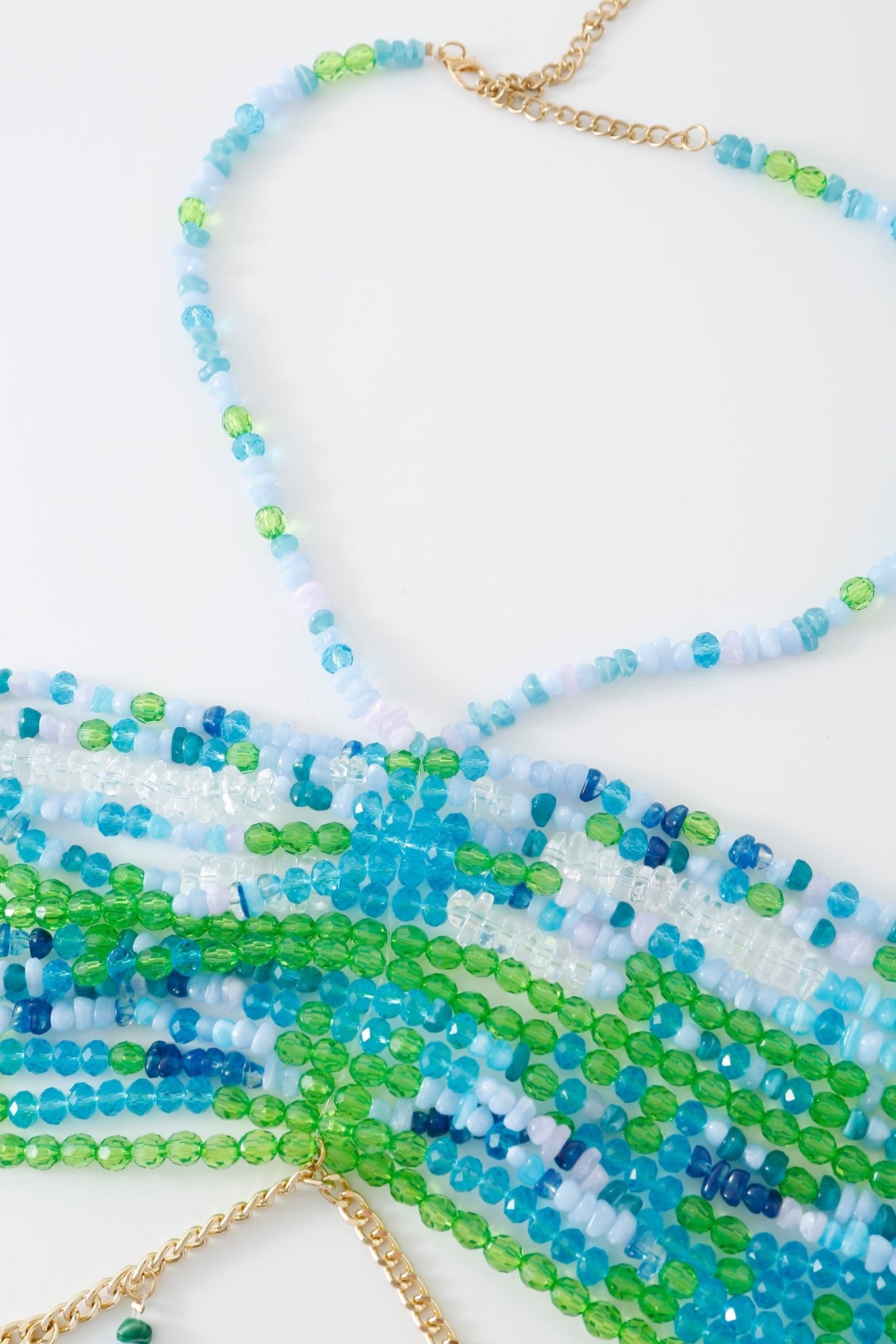 Close-up of the Lagoon w Pashmina- Full Set—a beaded necklace with blue, green, and clear beads, featuring a gold chain and clasp—arranged on a white surface. This elegant accessory pairs beautifully with a beaded top or adds a subtle shimmer to your look beneath a fringe pashmina wrap.