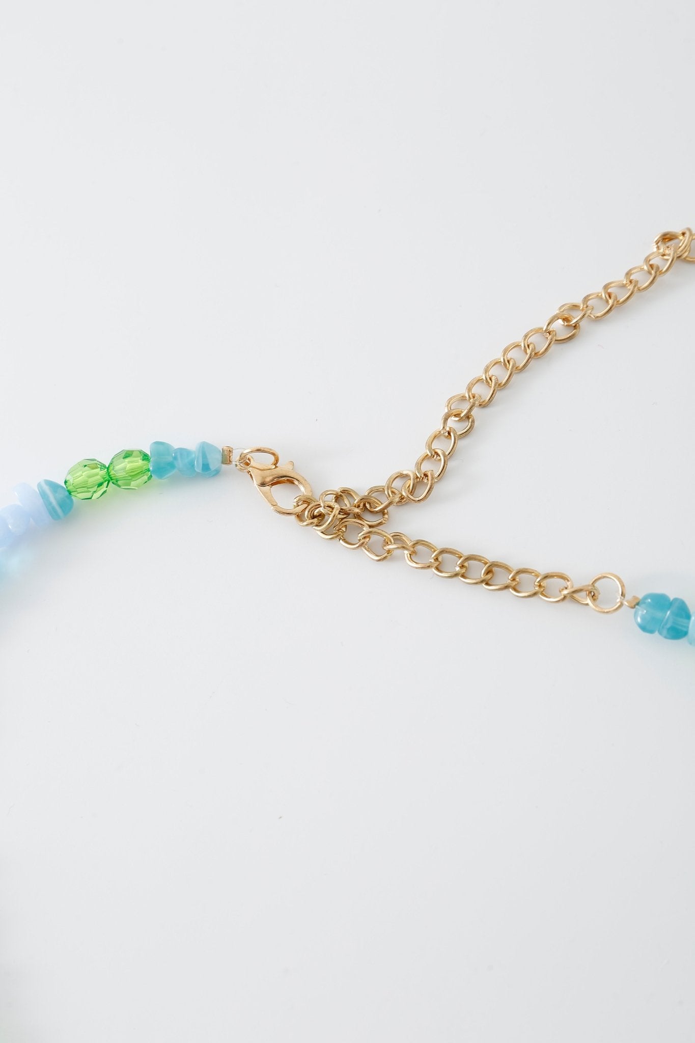 Close-up of the Lagoon w Pashmina- Full Set: a gold chain necklace with a lobster clasp, featuring blue and green beads on one side, placed against a white background—perfect for pairing with a beaded top or your favorite Lagoon lingerie.