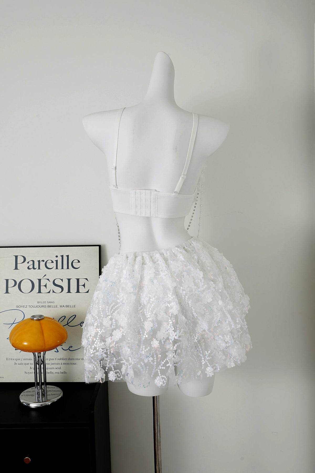 A white mannequin displays a white lace skirt and bra. A framed "Pareil Poésie" print and an amber mushroom lamp sit on a black table beside it.
