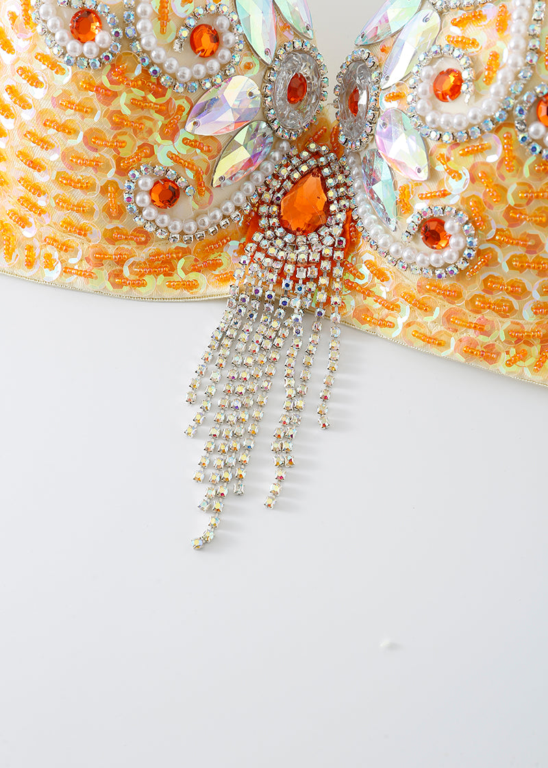 Close-up of the Lustre - Coral - Top, an ornate costume bra embellished with orange gems, iridescent sequins, pearls, and sparkling fringe, set against a yellow and orange fabric background.