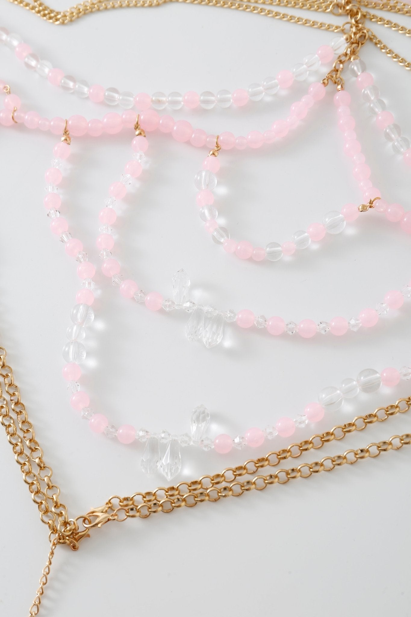 The Rosette w Pashmina - Full Set features gold chains and beaded necklaces with pink and clear beads, elegantly arranged on a white surface to create a dazzling ensemble that would beautifully complement your favorite fringe pashmina wrap or delicate beaded top.