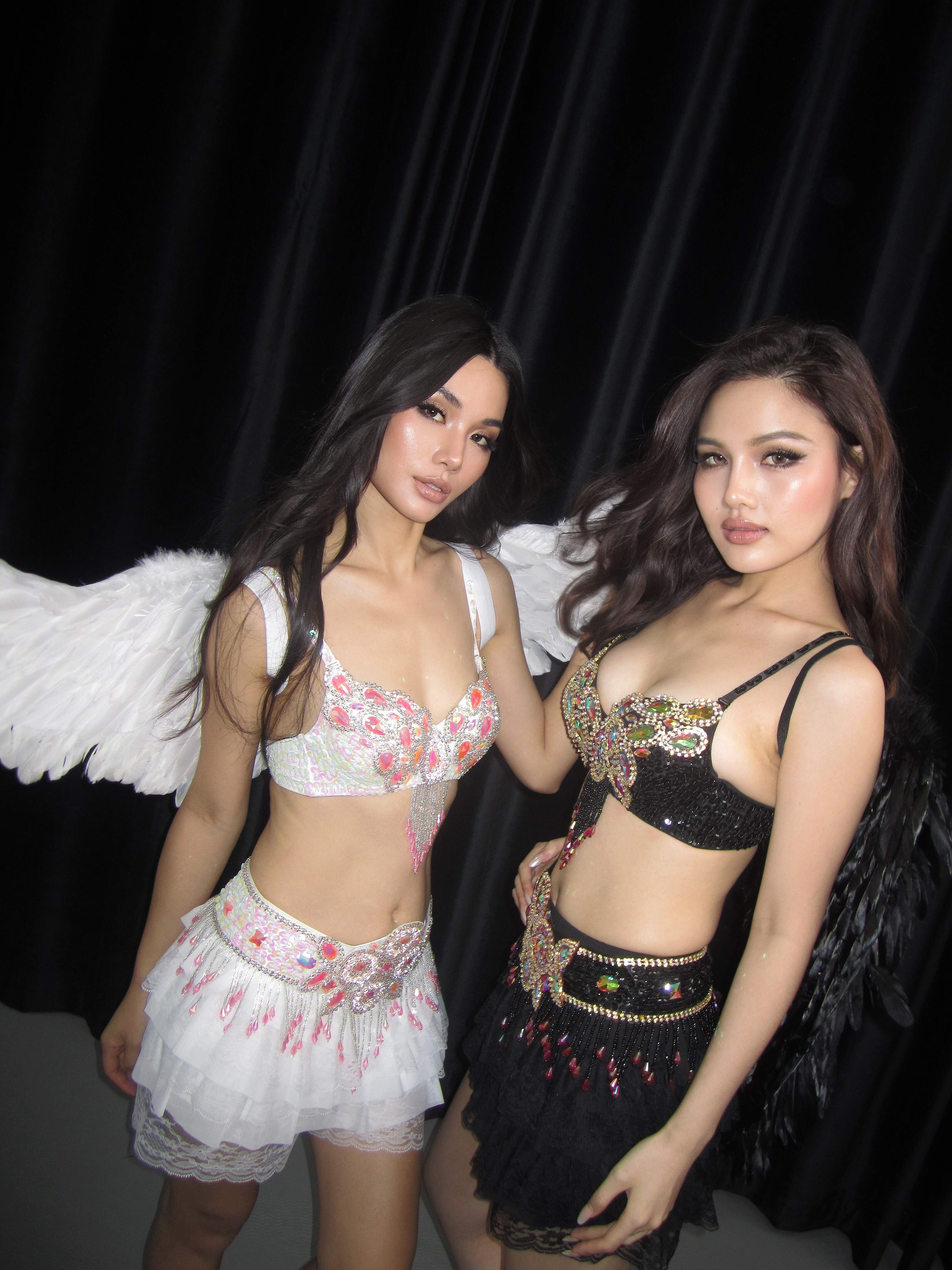 Two women stand side by side in front of a black curtain, each showcasing the perfect rave or festival look—one wearing the Vixen Full Set in White with angel wings, while the other complements her in black.