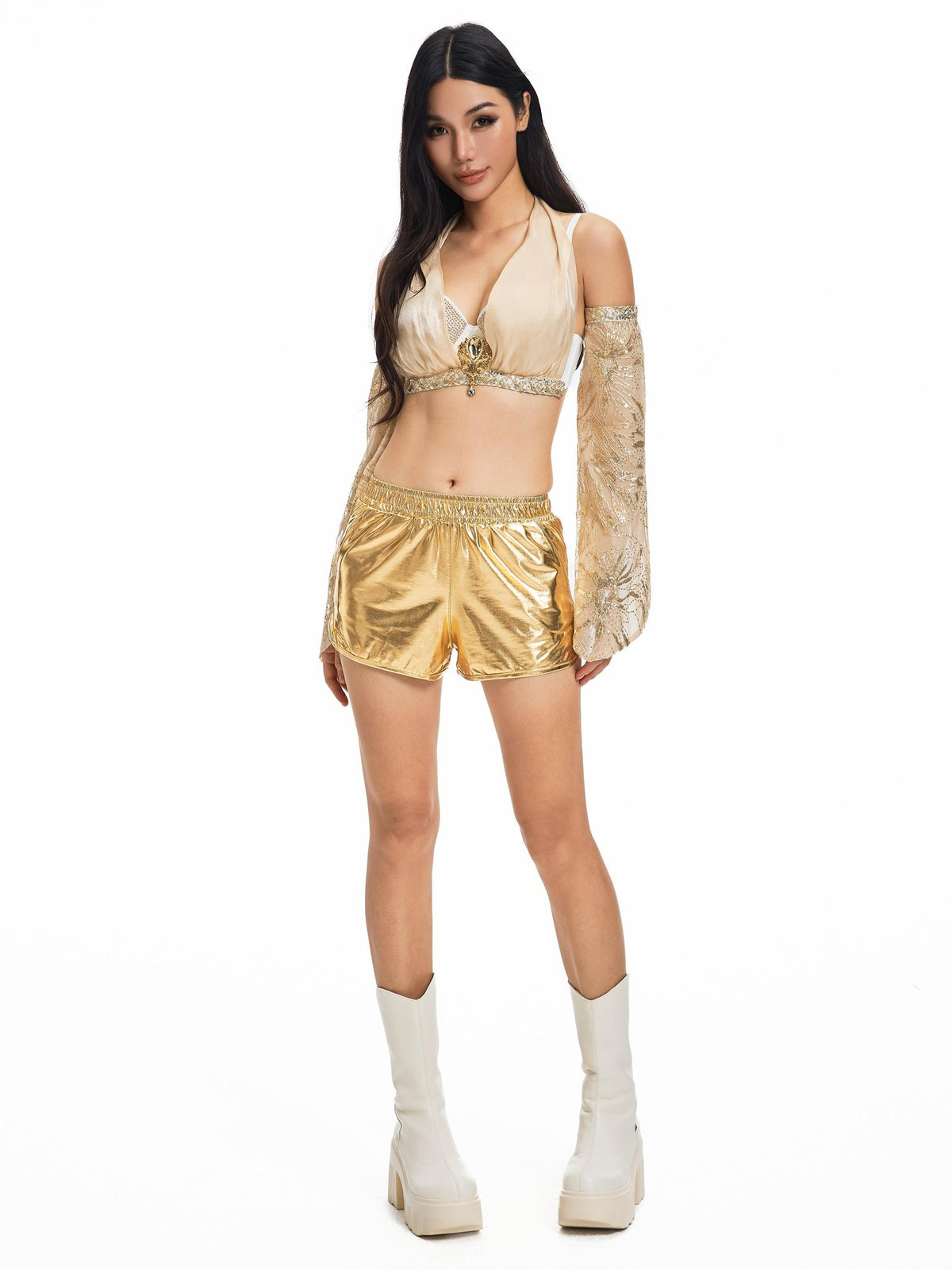 A woman models the Selene-Full Set-Gold, a stunning festival outfit consisting of a gold crop top with crystal details, matching gold shorts, sheer gold sleeve, and white platform boots, standing against a white background.