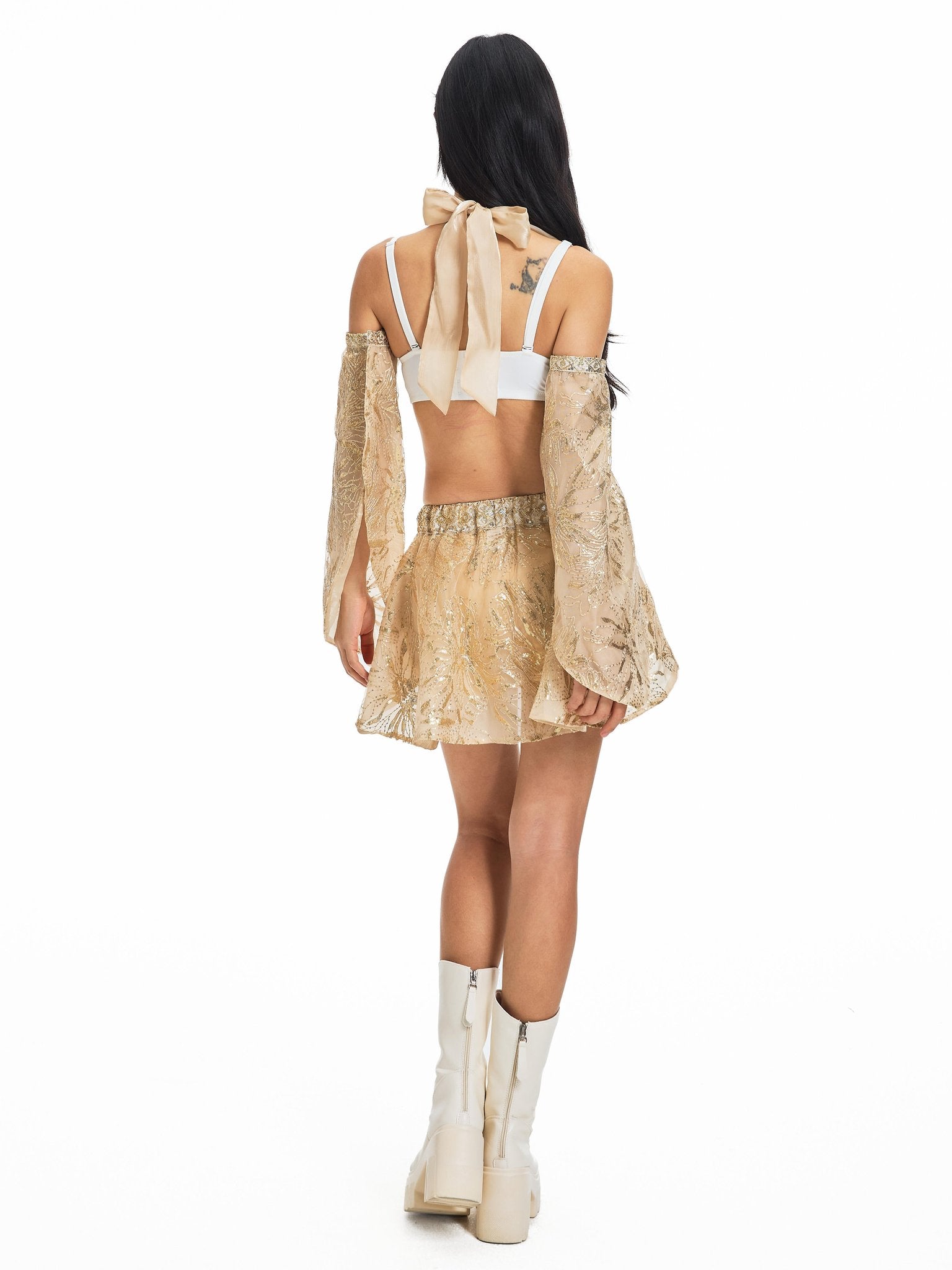 A woman faces away, wearing the Selene-Full Set-Gold—a striking gold skirt and coordinating long-sleeve open-back top, styled with a white bralette, large cream bow, and white knee-high boots—creating an effortlessly stylish festival look against a plain white background.