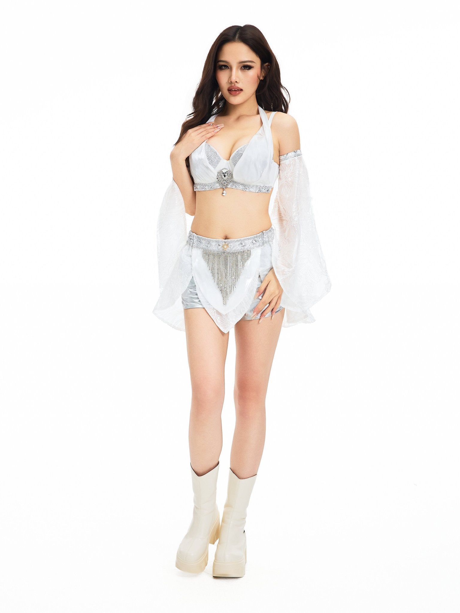 Dressed in the Selene-Full Set-White, she stands against a white background, radiating style in this dazzling four-piece ensemble featuring bell sleeves, shorts, and paired with platform boots—a striking festival outfit designed to make a statement.