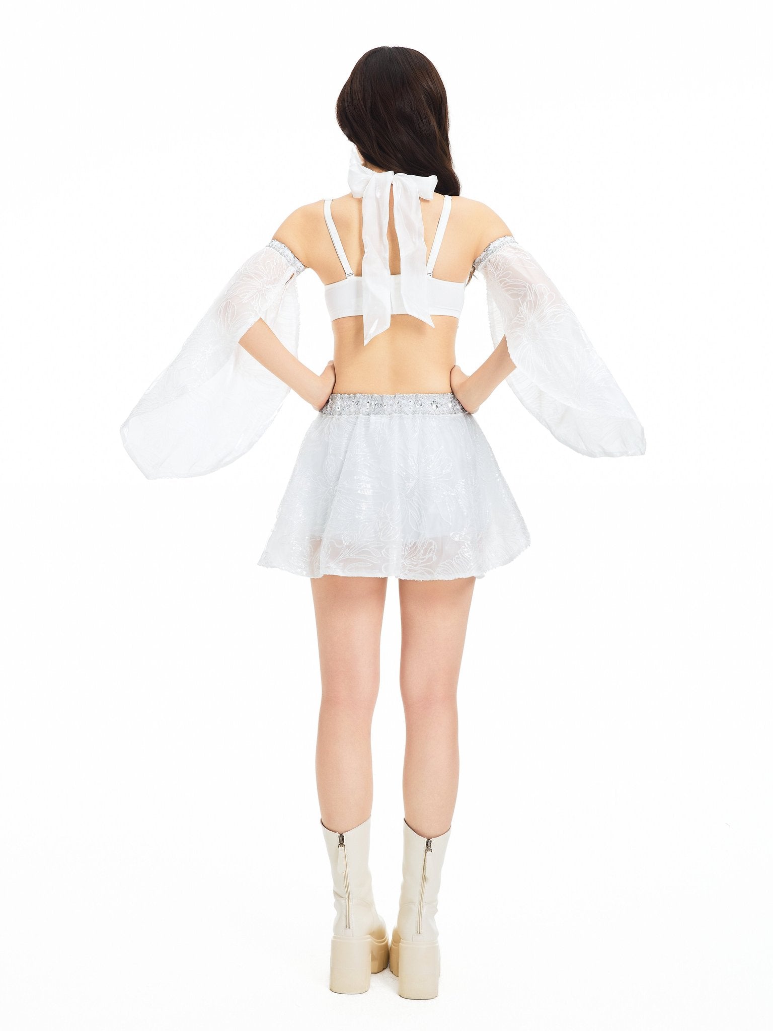 A person stands facing away against a plain white background, wearing the Selene-Full Set- White: a goddess-inspired ensemble featuring a white crop top with puffed sleeves, a short white skirt, and knee-high platform boots.
