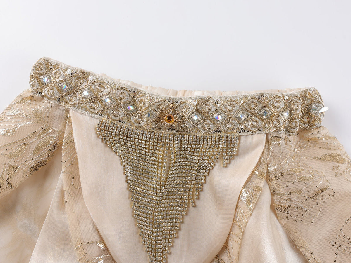 A close-up of the Selene-Full Set-Gold showcases an ornate, cream-colored fabric adorned with intricate gold embroidery, beadwork, and sequins in a decorative triangular pattern, elegantly finished with shimmering rhinestone fringe—perfect for elevating any festival outfit.